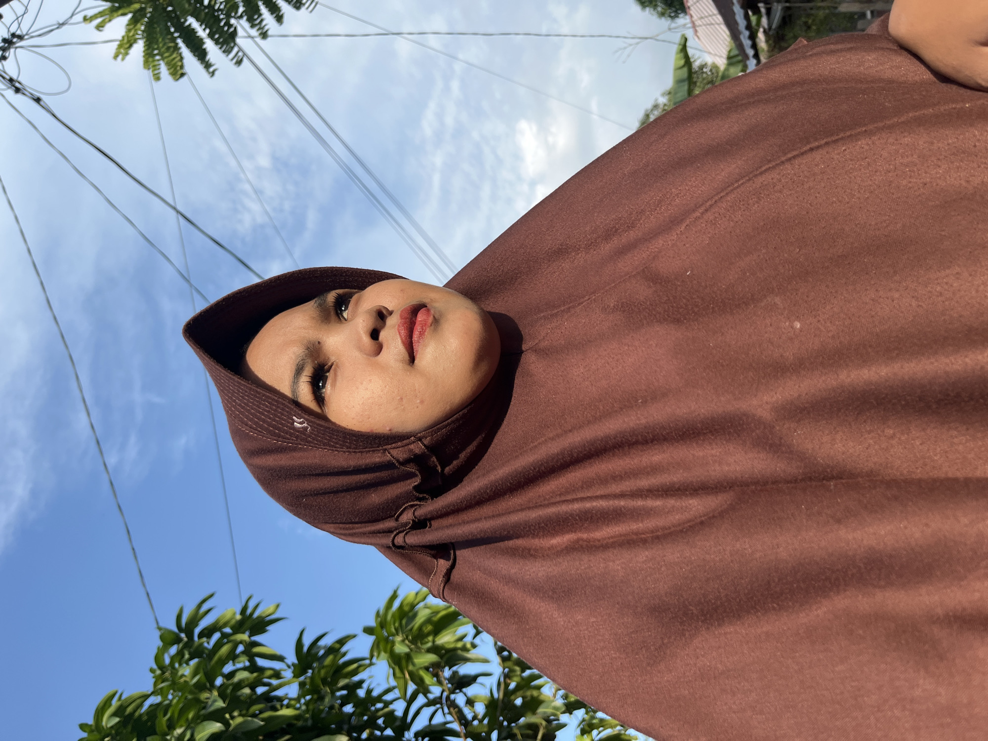 A medium shot captures a young woman wearing a dark brown hijab, looking upwards and to the right. Her face is turned slightly away from the camera, with her eyes gazing towards the sky. She has fair skin and is wearing lipstick. The hijab is draped around her head and shoulders, with subtle ruffles along the edge. In the background, a bright blue sky with wispy clouds is visible, along with power lines and some foliage, including a tree on the left and palm fronds on the right. A glimpse of a tiled roof and houses is also present in the far background. The lighting suggests it is daytime, with sunlight illuminating the scene. The image appears to be taken outdoors in what could be a residential area.