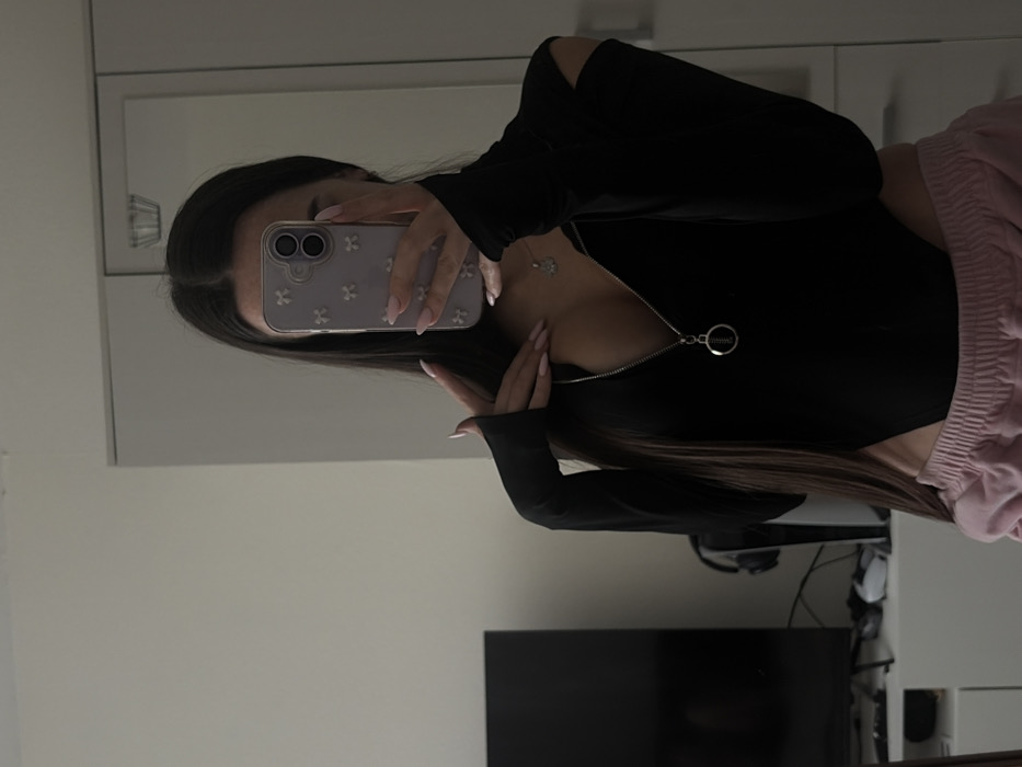 An indoor scene in what appears to be a residential room in the city of Krasnaya Zarya, Russia, is depicted. A single individual, a female, is visible from the chest down, reflecting in a mirror while holding a smartphone. She has long, dark hair parted in the middle. She is wearing a long-sleeved black bodysuit with an open zipper extending to below the chest, revealing a silver pendant worn around her neck. The bodysuit features thumbholes on the sleeves and cut-outs on the shoulders. Below the bodysuit, she wears light pink elastic-waist bottoms, possibly sweatpants. Her left hand is raised, with fingers splayed, near her collarbone. Her right hand holds a light purple smartphone with a bow-patterned case, obscuring her face and upper right chest. Her nails are manicured with light pink polish.
The background, visible around the individual's reflection, includes a light-colored wall on the left. On the far left, a dark-colored flat-screen television is positioned on a white console or cabinet. To the right of the television, a white gaming console (possibly a PlayStation 5) stands upright, with dark headphones resting near its base. Further to the right, a large, light-colored wardrobe or cabinet with reflective surfaces is visible, suggesting the mirror is integrated or placed against it.