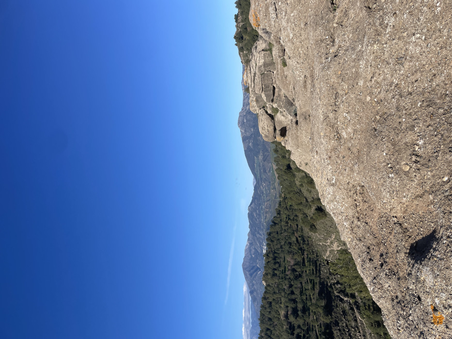 The image shows a view from a cliff overlooking a valley. The cliff is made of rough, light brown rock. The valley is covered in dense vegetation, with trees and shrubs. In the distance, mountains can be seen. The sky is a clear, bright blue. The image was taken in Álora, Spain, at the coordinates 36.827° N, 4.722° W. No people are present in the image.
