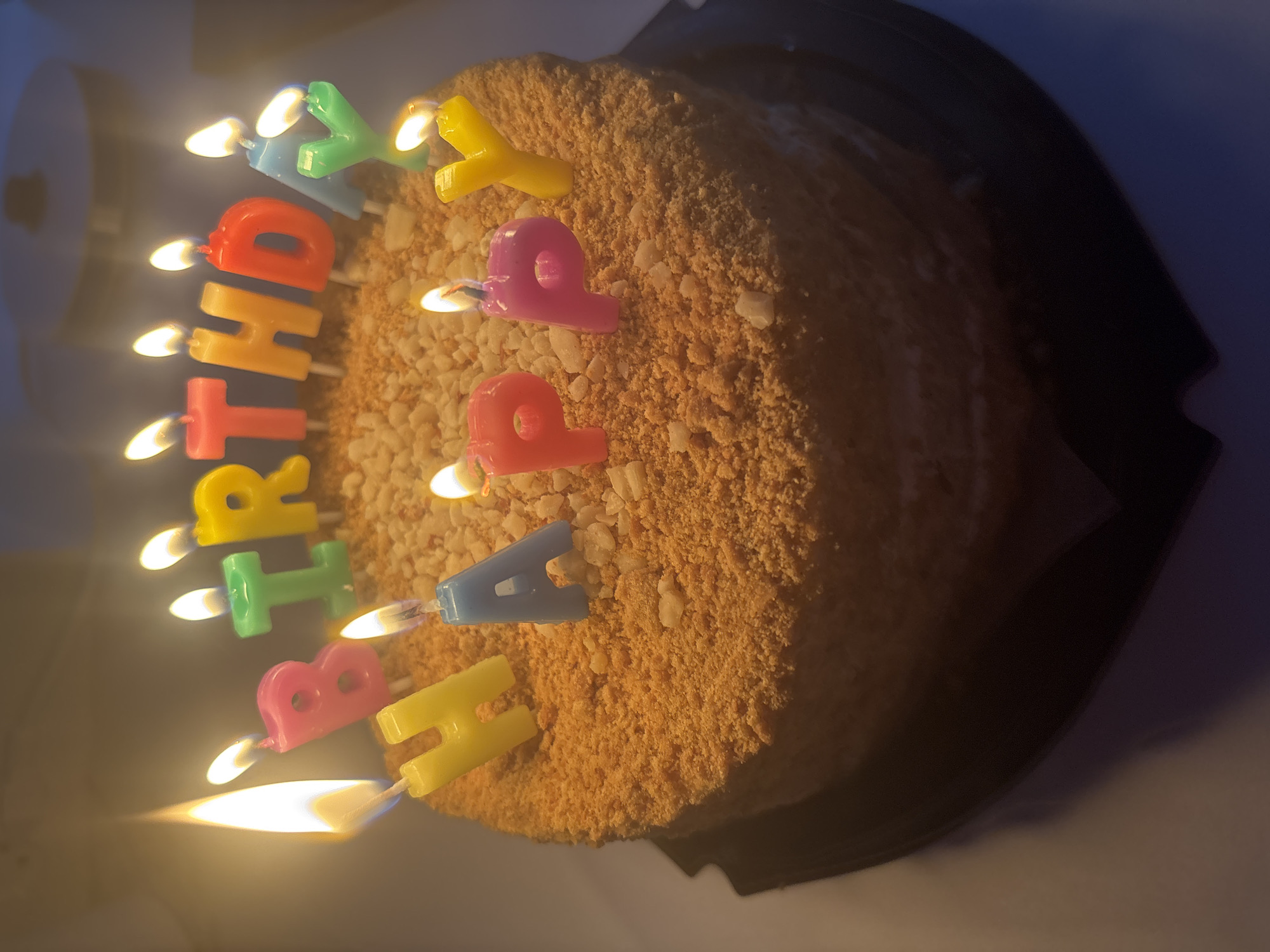 A lit birthday cake is shown from a slightly elevated perspective. The round cake is covered in a tan, crumbly material, possibly cake crumbs or nuts. Letter-shaped candles that spell "HAPPY BIRTHDAY" are stuck into the cake's surface; they are lit, with visible flames. The letters are various colors including yellow, red, pink, blue, and green. The cake sits on a dark-colored, likely plastic, circular tray. The background is dimly lit and blurred, suggesting a room interior, but is out of focus and lacks detail. There is nothing in the image to indicate a location in the city of Chaikovsky, Russia. The focus is primarily on the cake and the lit candles.