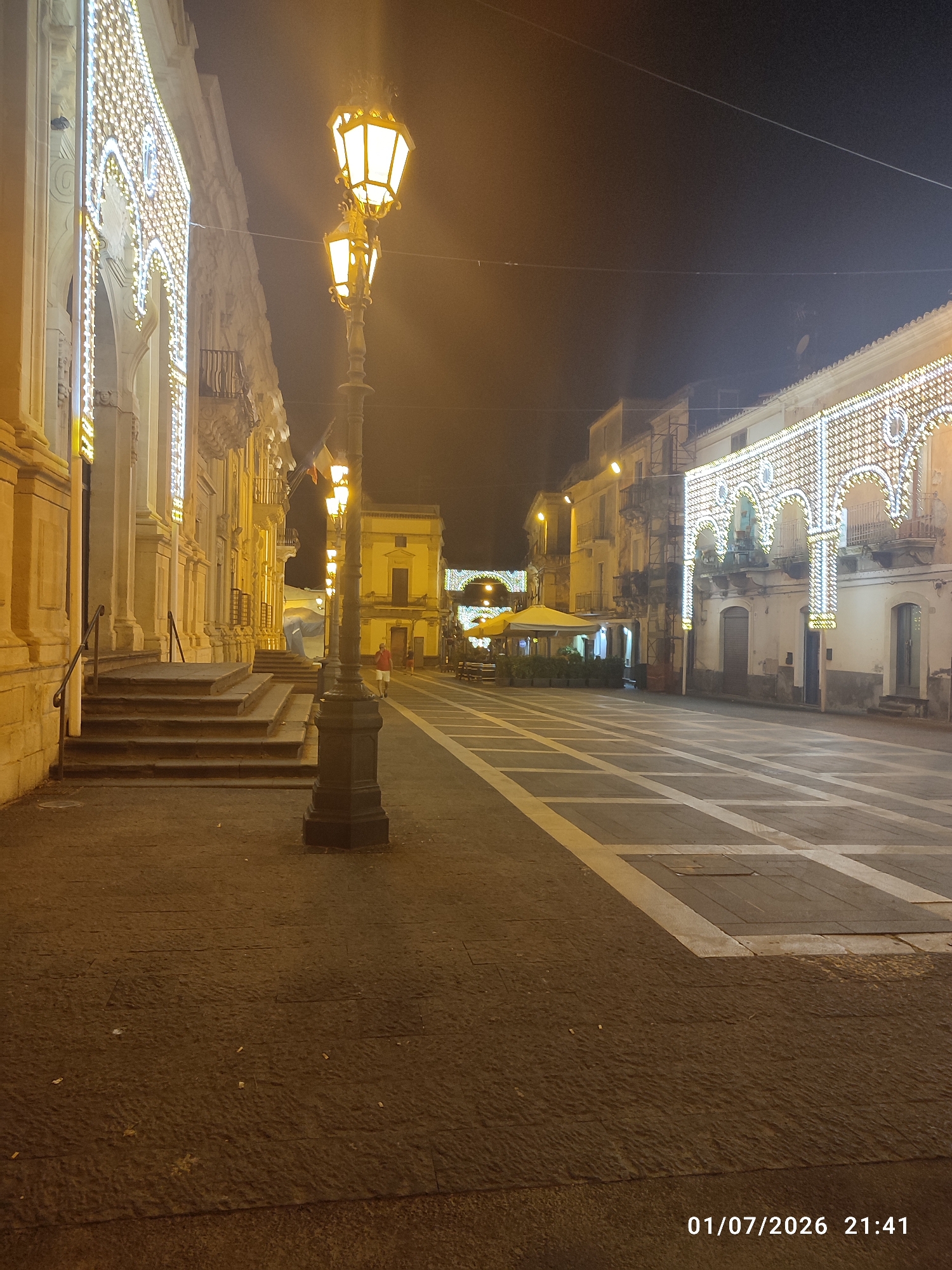 The image depicts a nighttime scene in a city square in Militello in Val di Catania, Italy. On the left, a building facade is decorated with intricate, illuminated patterns, possibly a historic building or a church. Adjacent to this building is a set of wide, stone stairs with handrails. A tall, dark lamppost with multiple lights stands in the foreground, illuminating the paved square. The square itself has a patterned design of light-colored squares on a darker paved surface. In the background, other buildings line the square, some also adorned with decorative lights. There are outdoor seating areas with umbrellas, suggesting a cafe or restaurant. One person, wearing a red shirt and white shorts, is visible walking away from the viewer on the right side of the square. Another person is partially visible near the outdoor seating area. The sky is dark, indicating nighttime. A timestamp "01/07/2026 21:41" is present in the lower right corner of the image.