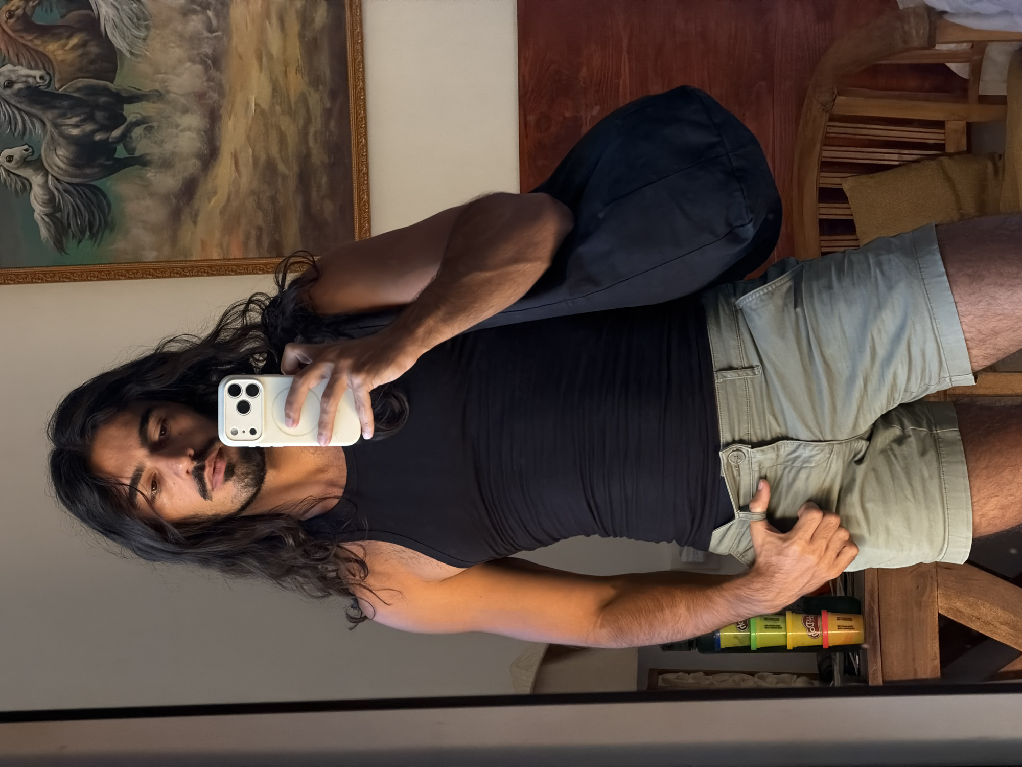 A man with long, dark curly hair and a mustache is taking a selfie in a mirror. He is wearing a black tank top and light green shorts. He is holding a white iPhone with a MagSafe charger attached. A black bag is draped over his shoulder. In the background, there is a painting of horses, a red wooden wall, and a wooden chair. Play-Doh containers are visible on a shelf or surface below the chair. The photograph appears to be taken indoors in North Kuta, Indonesia.