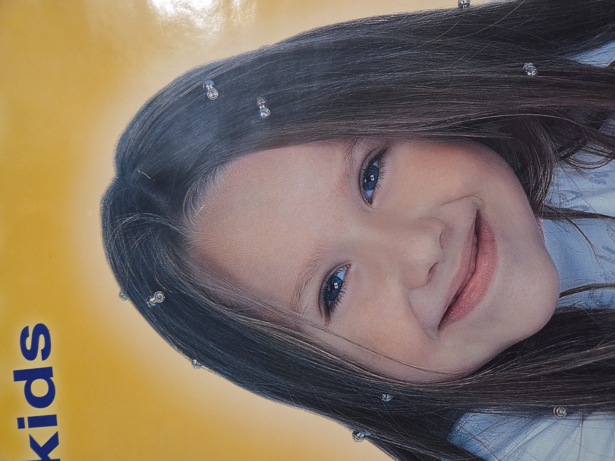 A close-up, slightly angled image features a young girl with shoulder-length, dark brown hair adorned with small, decorative hair accessories. The girl has fair skin, blue eyes, and a gentle smile. The background is a yellow and blue color. The visible text "kids" is in blue. The image appears to be a print or advertisement. No specific location in Pleven, Bulgaria, is evident from the photograph.