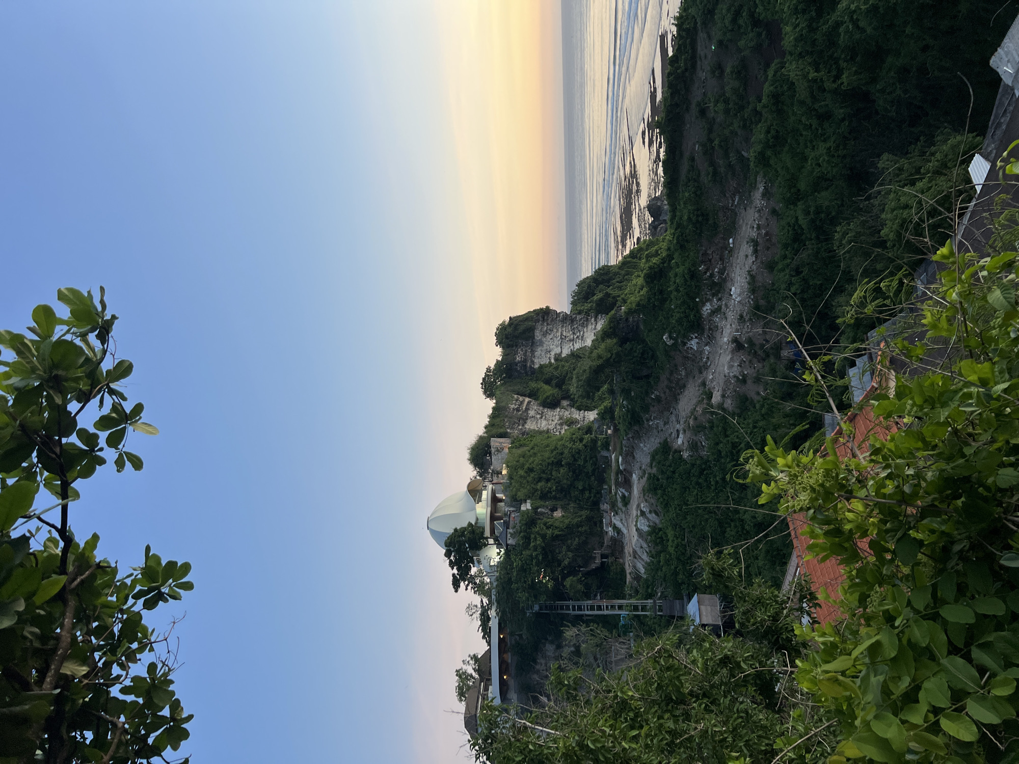 Here is a forensic description of the image.
The image depicts a coastal scene, likely taken from an elevated position. The foreground shows vegetation, including trees with broad leaves. The middle ground features a cliff face covered in greenery. A structure with a dome-shaped roof is visible on the cliff, along with what appears to be a walkway or bridge extending from it. The background reveals the ocean under a sky transitioning from blue to orange, suggesting either sunrise or sunset. The location is Unknown, Unknown.
