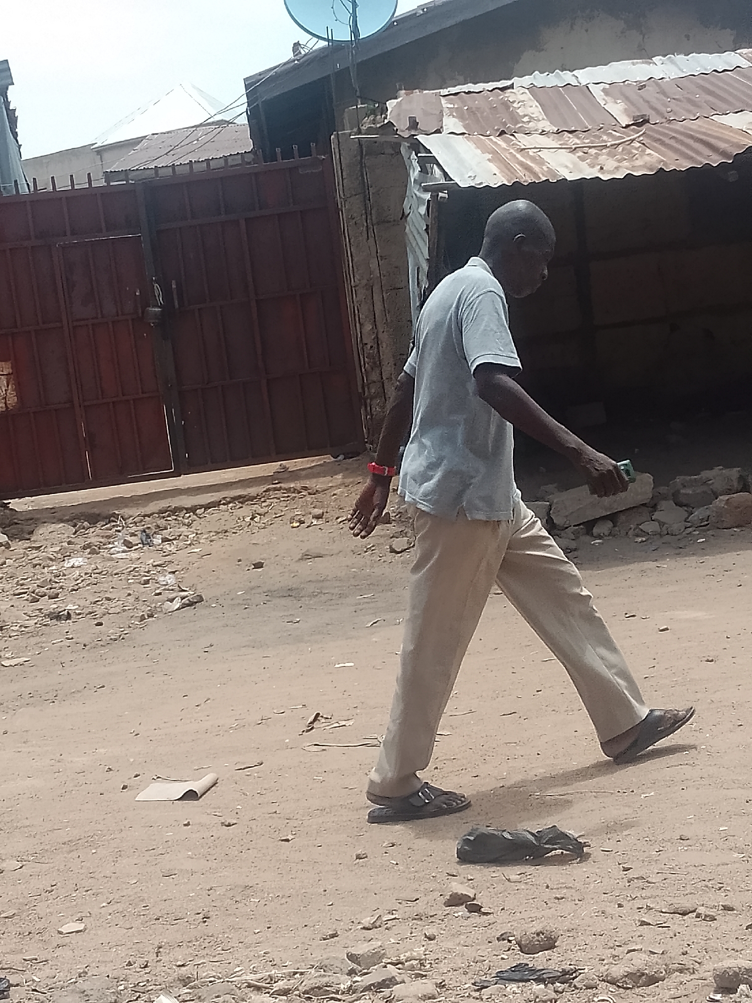 A man is walking in the city of Pantame, Nigeria. The man is wearing a grey t-shirt, tan pants, and sandals. He is holding a small object in his right hand and has a red watch on his left wrist. In the background, there is a gate on the left and a building with a corrugated metal roof to the right. The ground appears to be sandy and littered with debris. There is a piece of paper and a black plastic object lying on the ground.