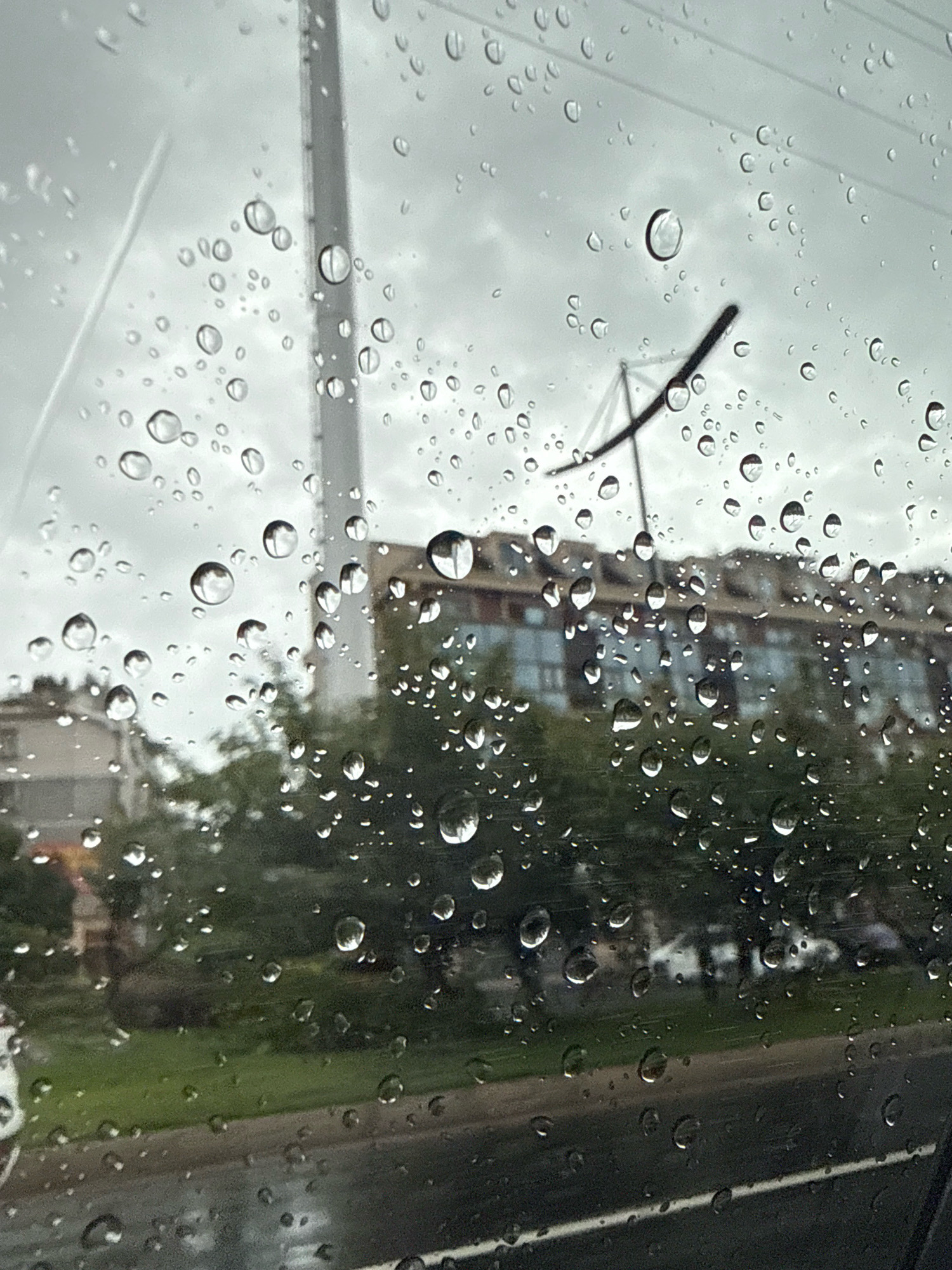 A close-up, eye-level, exterior shot captures a rain-streaked view through a window, likely from inside a vehicle, looking out onto a cityscape. The foreground is dominated by numerous raindrops of varying sizes clinging to the glass, distorting the view of the background. In the mid-ground, a multi-story building with a light-colored facade and dark windows is visible, partially obscured by lush green trees. To the left, a light-colored residential building with multiple windows can be seen. A grey road with a white line marking the lane is visible in the lower portion of the frame. In the far background, the sky is overcast and a large, grey pole-like structure rises vertically, with a dark, angled object extending from it, possibly a piece of electrical infrastructure or a tower. The overall impression is one of a wet, urban environment, possibly during a downpour, on a cloudy day. The image does not provide specific identifiable landmarks or context to definitively place it in Yiwu, China.
