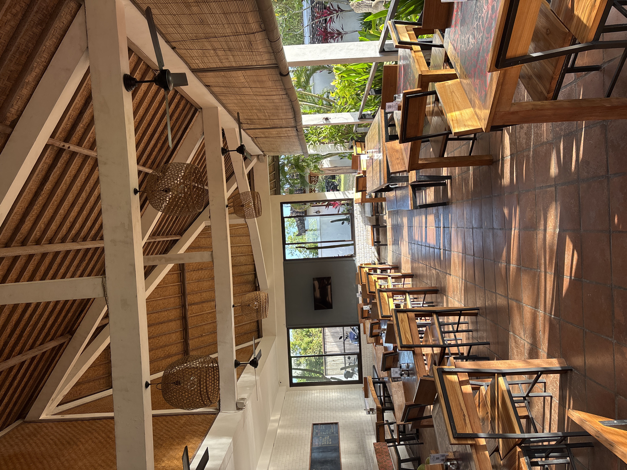 The image captures the interior of a restaurant in Kuta Selatan, Indonesia. The restaurant features a high, exposed roof structure with a thatched ceiling supported by white beams and columns. Wicker pendant lamps hang from the ceiling. The floor is tiled in a warm, reddish-brown color. Tables and chairs are arranged in rows, primarily made of wood with dark metal frames. Sunlight streams into the restaurant, creating strong shadows on the floor and furniture. The restaurant's exterior is visible through large windows, showing lush greenery. There are no people visible in the image.