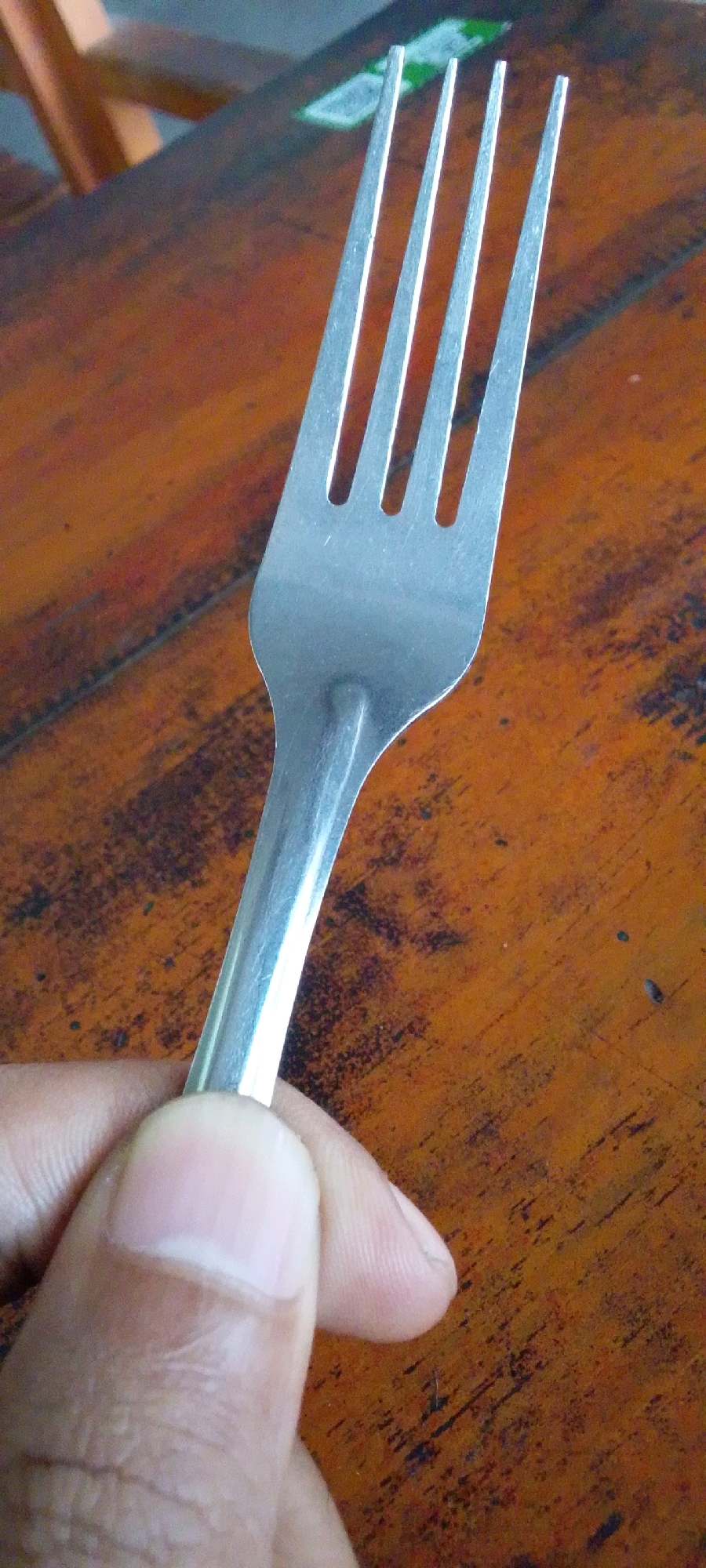 A close-up shot shows a person's thumb and forefinger holding the handle of a metal fork. The fork is positioned over a wooden table with a reddish-brown, textured surface. The table appears to be outdoors, possibly in Ngaglik, Indonesia, given the lighting and casual setting. The background is blurred, but hints of a wooden chair and a green object, possibly a label or packaging, are visible. The focus is on the fork and the hand holding it, suggesting a moment of pause or preparation before a meal.