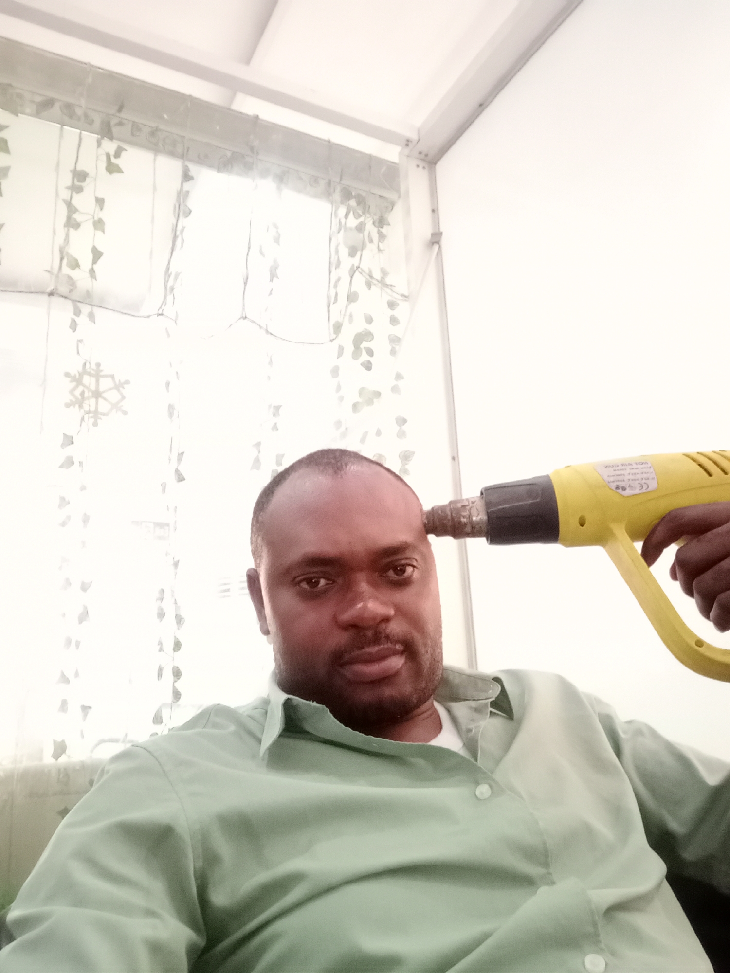 A dark-skinned man is captured in a medium close-up shot, seated indoors in Gwarinpa, Nigeria. He has short dark hair and a short beard, and wears a light green collared shirt over a white undershirt. He looks directly at the camera with a neutral expression. With his right hand, he holds a bright yellow heat gun, pressing its nozzle to his right temple in a suggestive, possibly humorous, pose.
The background features a window covered by sheer white curtains adorned with light grey leaf patterns and a visible snowflake decoration. Diffused natural light filters through the curtains. Above the curtains, light-colored structural elements, possibly window frames or ceiling beams, are visible against a white wall and ceiling.
Visible text on a white sticker on the side of the yellow heat gun includes "HOT AIR TOOL" and other smaller, indistinct text that may contain certification marks. The bright lighting indicates it is daytime. The man's calm demeanor, despite the unusual action, suggests a playful or symbolic intent behind the pose.
