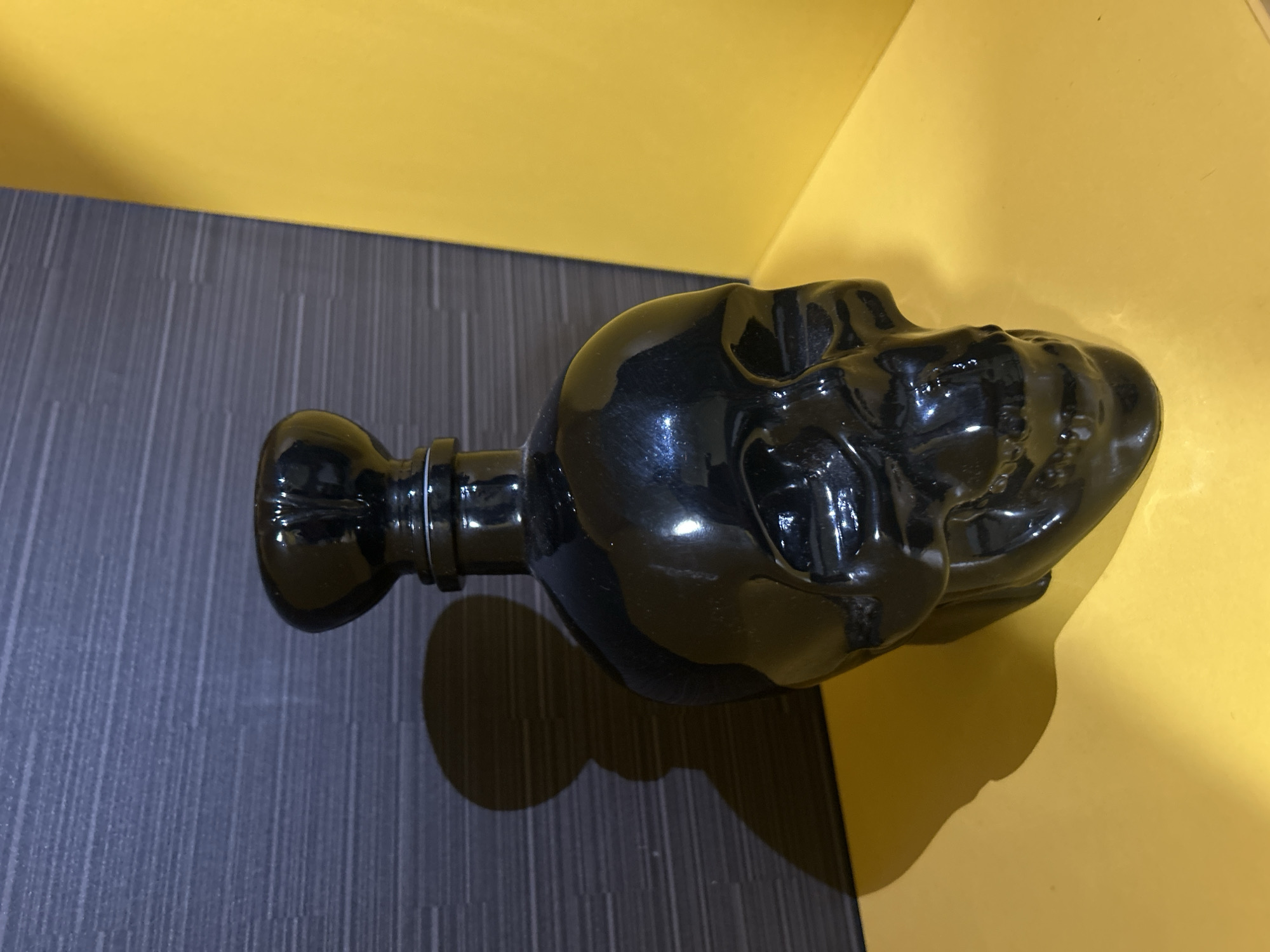 The image shows a black, skull-shaped bottle lying horizontally on a surface. The surface appears to be composed of two sections: a yellow section and a gray, lined section. The bottle's shadow falls onto both surfaces. The bottle is glossy, reflecting light. There is no identifiable location in Varna, Bulgaria, nor are there any people depicted in the image.