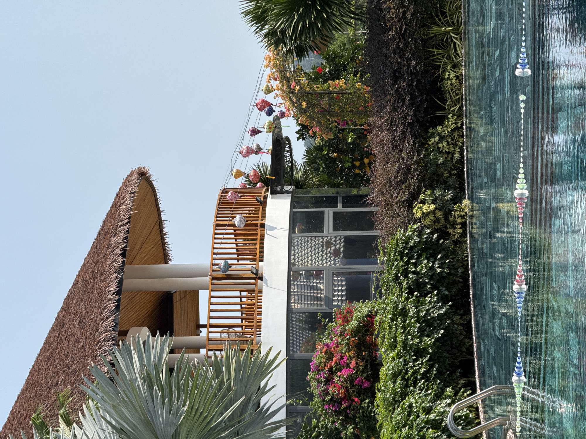 The image depicts an architectural composition in Phú Quốc, Vietnam. The left side features a thatched roof structure supported by cylindrical columns. A wooden ladder-like structure ascends the side of a white building with glass windows. Above, a trellis with floral decorations and lanterns is visible. Dense vegetation, including various plants and flowers, fills the scene. To the right, a glass waterfall feature with decorative glass beads and a stainless steel handrail is present. The sky is light blue.