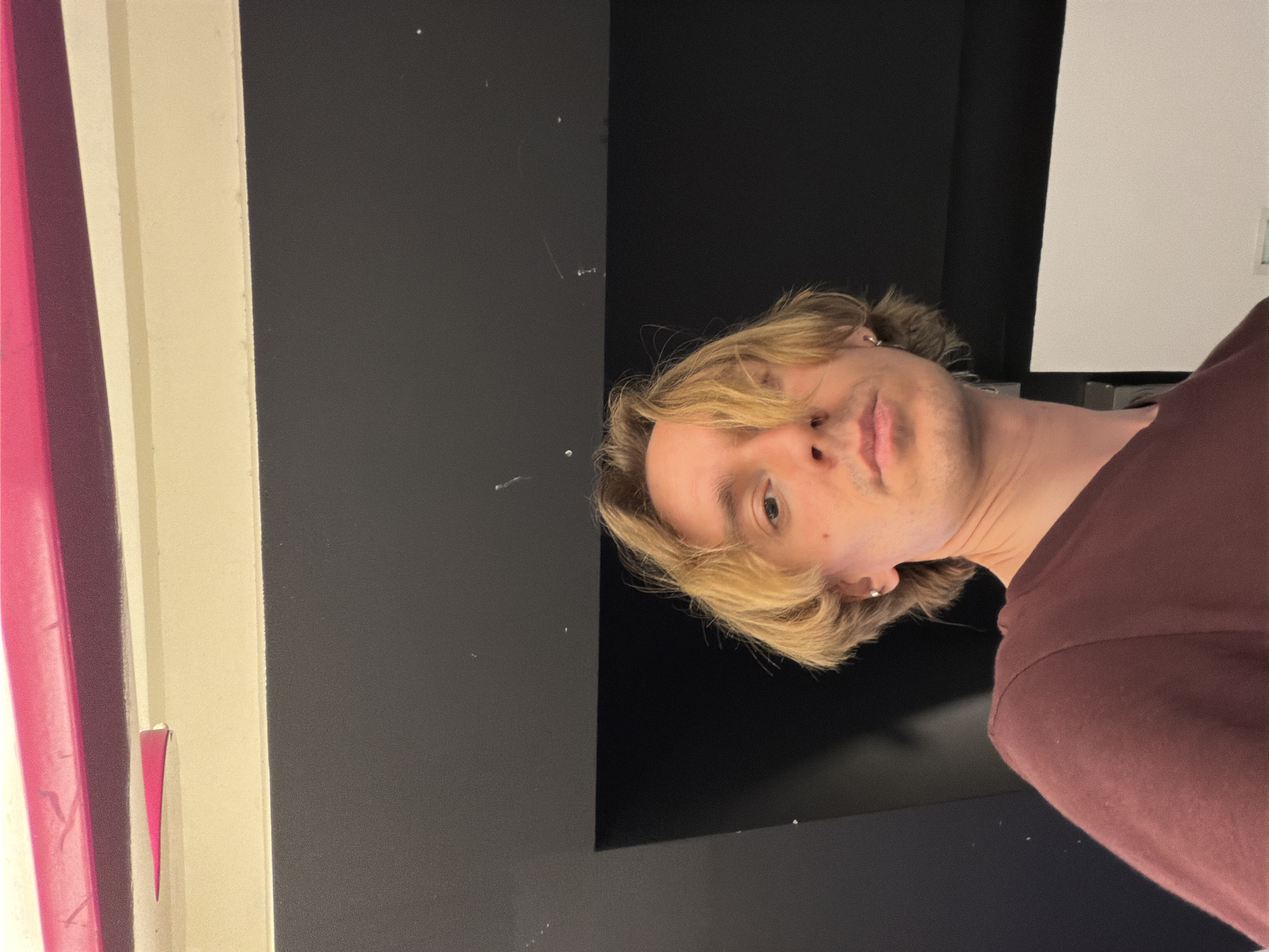A photograph depicts a blond-haired individual wearing a dark red shirt, positioned against a backdrop of contrasting dark gray and white panels. No information in the image allows for inferring the location as Budva, Montenegro. The person is partially framed by the dark gray and black section, with a glimpse of what appears to be architectural details – including a pink stripe, a cream-colored stripe, and a gray stripe – visible to the left of the dark gray panel. The white panel is located on the upper right part of the image. The focus is on the individual's face, which is turned towards the camera. The person's head is tilted to the left, with the lower right area of the image occupied by the dark red shirt.