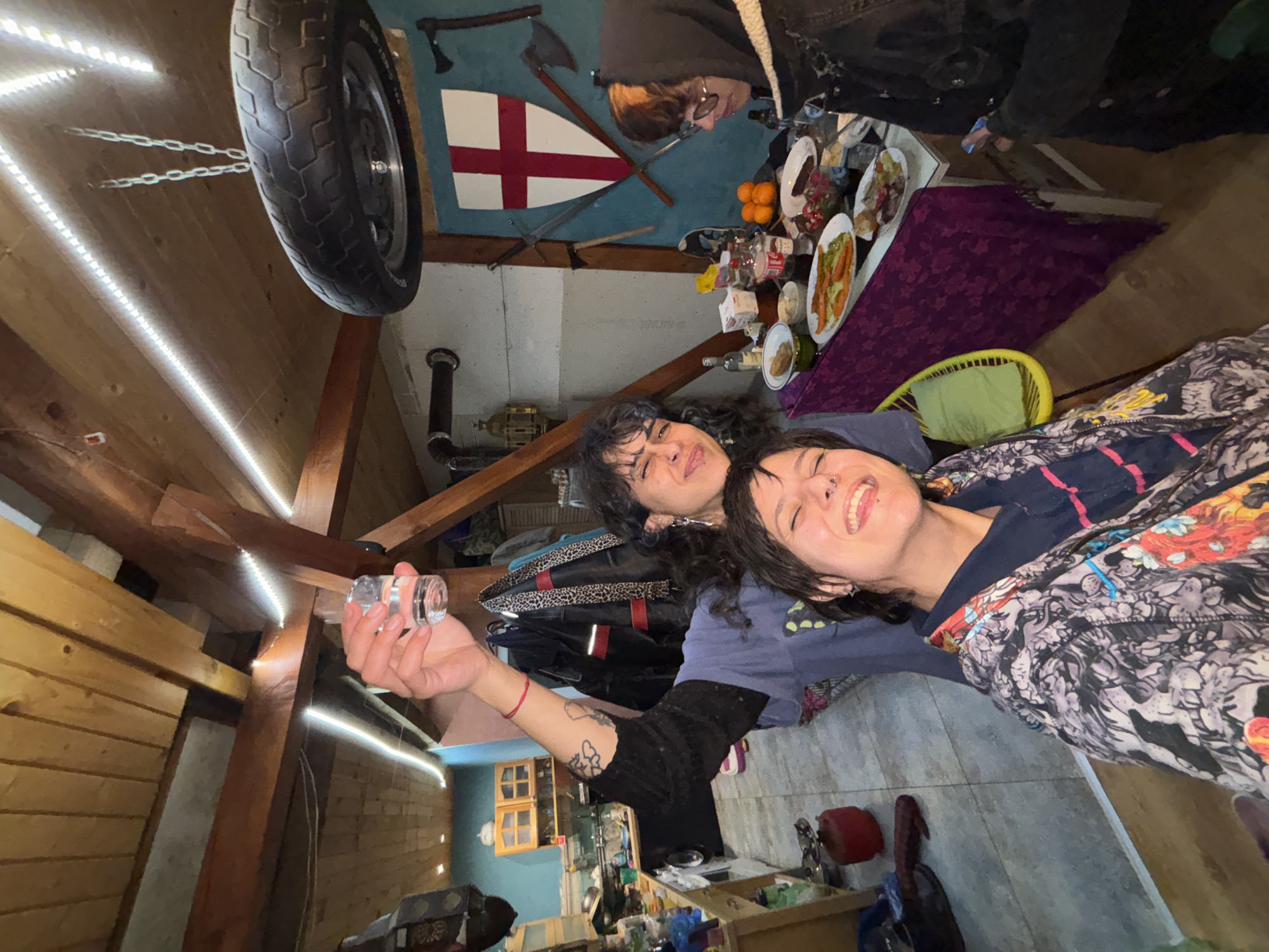The scene appears to be indoors, likely in a converted attic space. Two individuals, both female, are the primary focus. They are close together and appear to be interacting with each other, with one holding up a small glass container. The environment is cluttered and eclectic, with a motorcycle tire suspended from the ceiling by chains, and decorative items like axes and a shield painted with a red cross on the wall. There is also a table laden with food and drinks. The location is unspecified; Verinsko, Bulgaria is not discernible from the image.