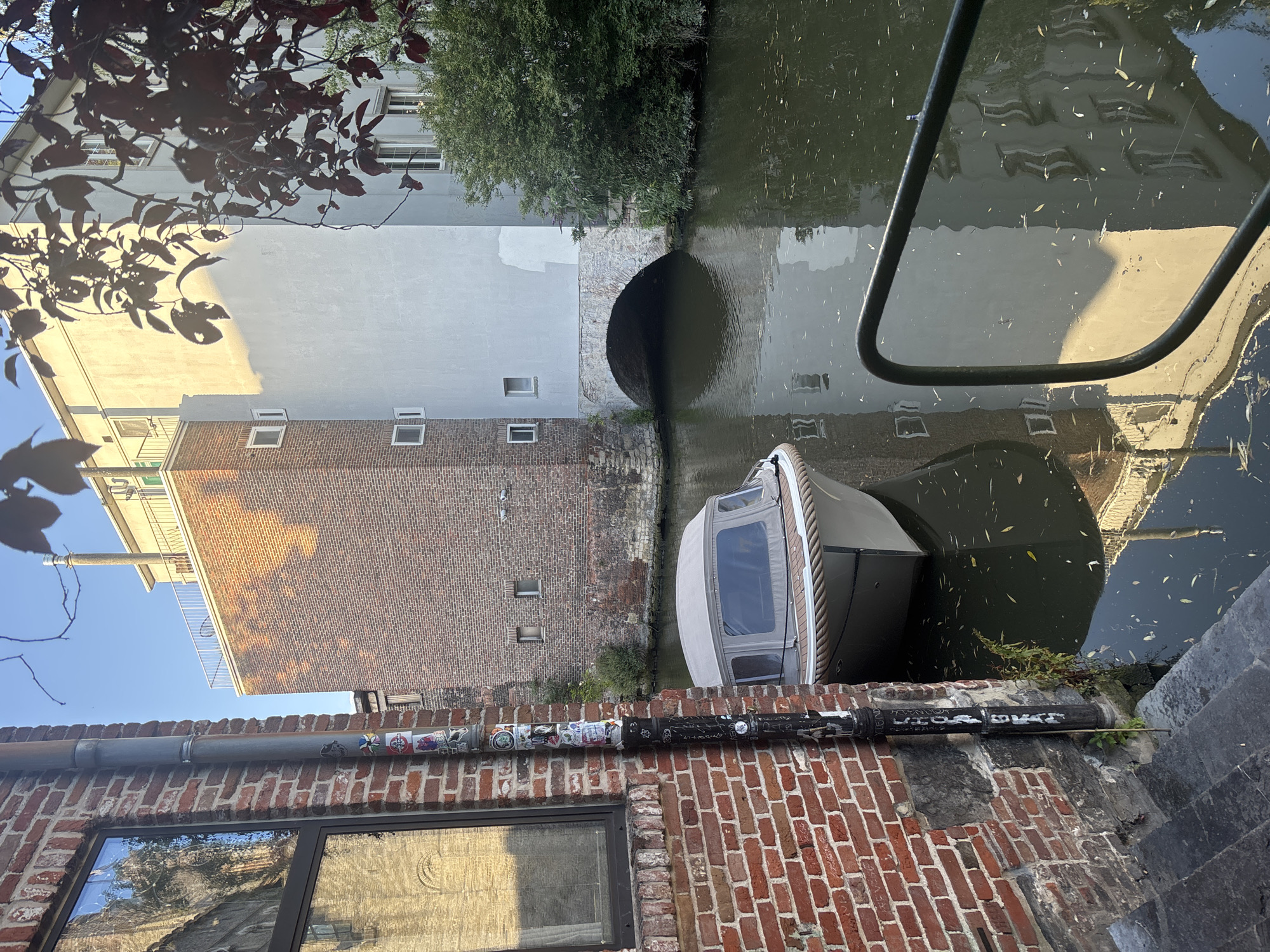 The image depicts a canal scene in Ghent, Belgium, likely in the historic city center. In the foreground, a portion of a red brick building with a large window is visible on the left, alongside a dark downspout covered in stickers. A small boat with a light-colored canopy is moored in the dark canal water, which reflects the surrounding architecture. The water also contains fallen leaves. Behind the boat, a stone archway leads into a building. The background features a multi-story brick building and a lighter-colored modern structure, with trees and foliage adding to the urban greenery. The sky is clear and blue, suggesting a sunny day.