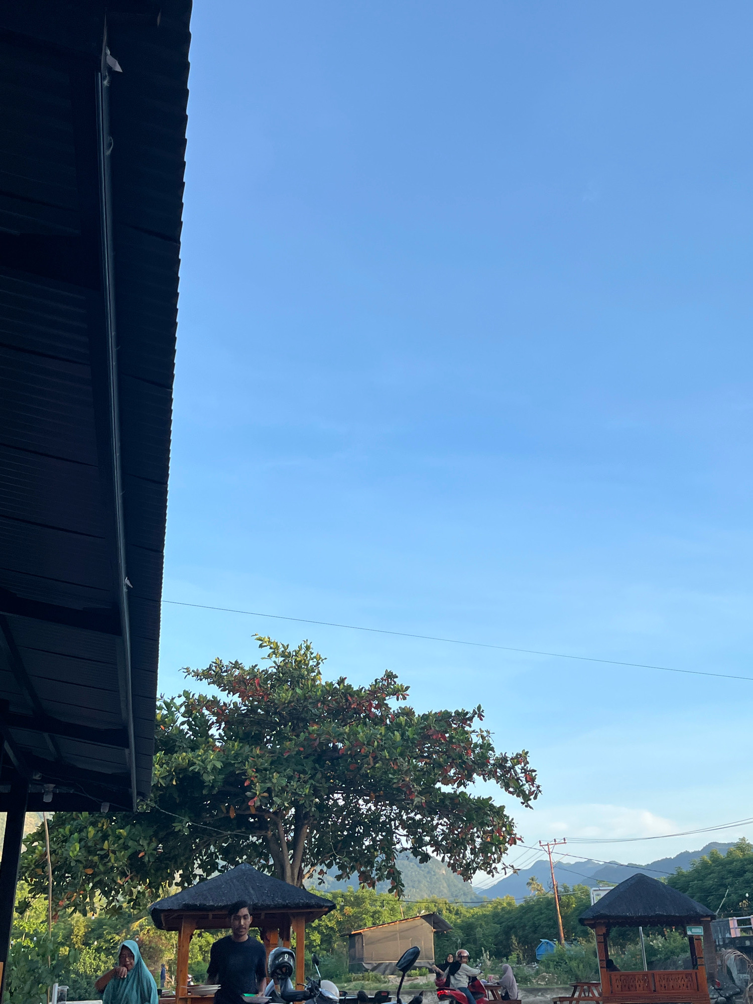 The image depicts an outdoor scene in Aneuk Paya, Indonesia, under a clear blue sky. In the foreground, a dark, corrugated metal roof juts in from the left side of the frame, casting a shadow. To the right of the roof, a lush green tree with red-tinged leaves is prominent. Below the tree, a small, dark-roofed wooden gazebo with ornate carvings stands on a raised platform. A man in a dark t-shirt is standing near the gazebo, holding a plate. Further to the right, another similar gazebo is visible. In the background, a line of mountains stretches across the horizon, interspersed with greenery and a utility pole. Several people are present, some sitting or standing around the gazebos, and two individuals are on a red motorcycle in the middle ground. The overall atmosphere appears to be casual and relaxed, possibly at a roadside eatery or rest stop.