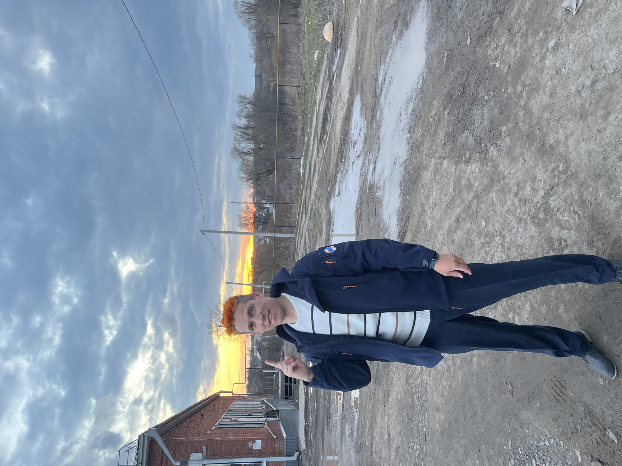 This image captures a man standing outdoors in Кварса, Russia, during what appears to be sunset or sunrise. The sky is a prominent feature, displaying dramatic, partly cloudy formations with a vibrant orange and yellow glow along the horizon where the sun is low, casting a warm light.
The central subject is a man with distinctive reddish-orange hair, standing with a slight smile or smirk and looking directly at the camera. He is dressed in a dark blue zip-up jacket, under which he wears a white sweater featuring horizontal black and brown stripes. He completes his attire with dark blue trousers and dark shoes. His right index finger is raised in a pointing gesture. A watch is visible on his left wrist, and a patch with a circular emblem is affixed to his left jacket sleeve.
The ground is an unpaved, muddy terrain with several large puddles, suggesting recent rain or poor drainage. To the left, a weathered brick building with windows is partially visible. In the mid-ground and background, a line of sparse, bare trees stretches across the landscape, indicative of a late autumn or winter season. Utility poles and overhead power lines are also present, adding to the semi-rural or industrial feel of the setting. There is no visible text in the image.