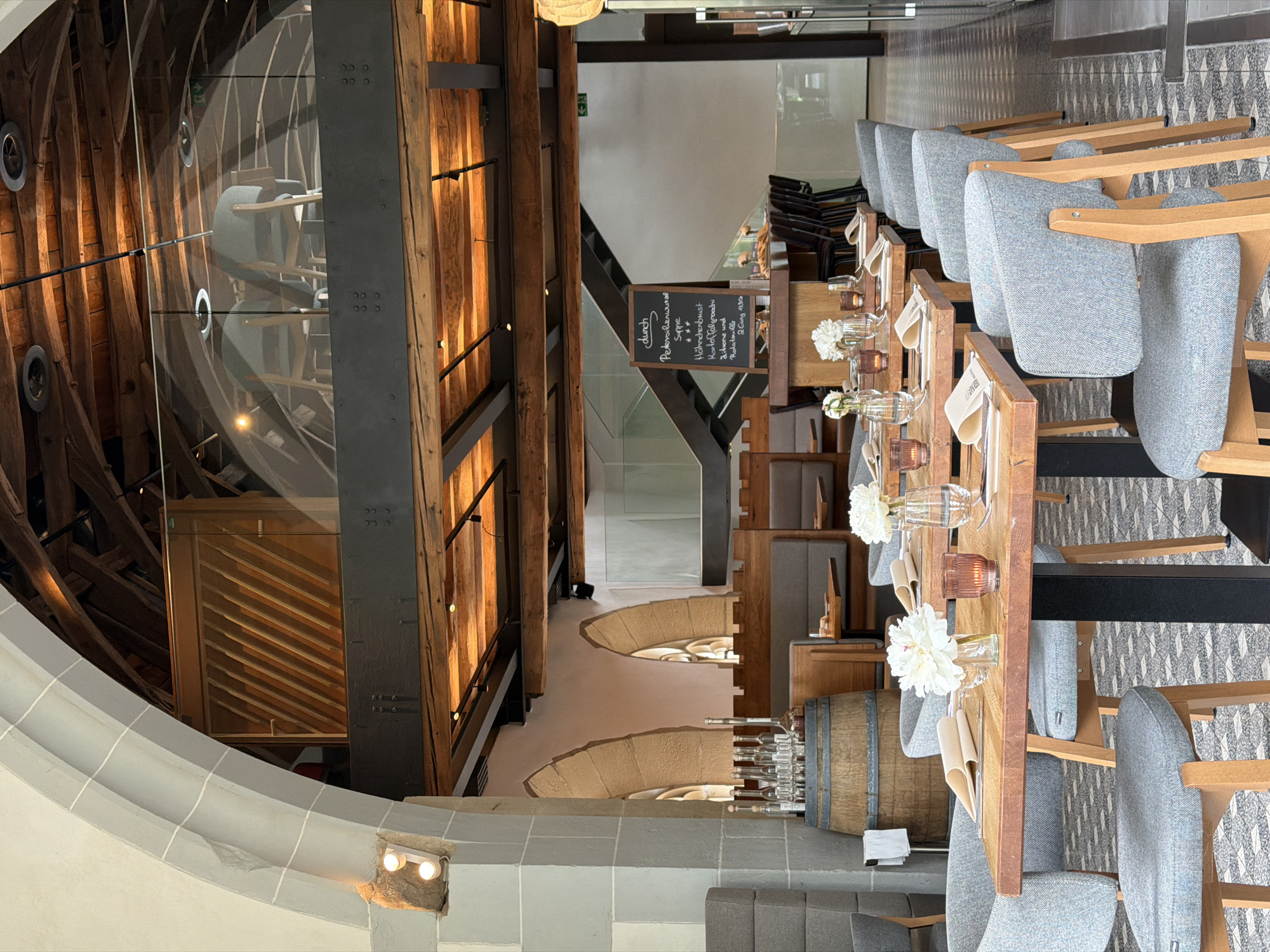 This image captures a restaurant interior in Bad Sobernheim, Germany. The building has a unique architectural style with exposed wooden beams and steel supports. There is a menu on a small blackboard displaying "Dunch" and a list of dishes. Tables are set with flowers, glassware, and napkins. Wooden chairs with gray fabric upholstery are arranged along the tables. A large wooden barrel is partially visible near a set of tables. Arched windows are seen in the background, suggesting a historical building. The overall lighting is warm, creating a pleasant ambiance.