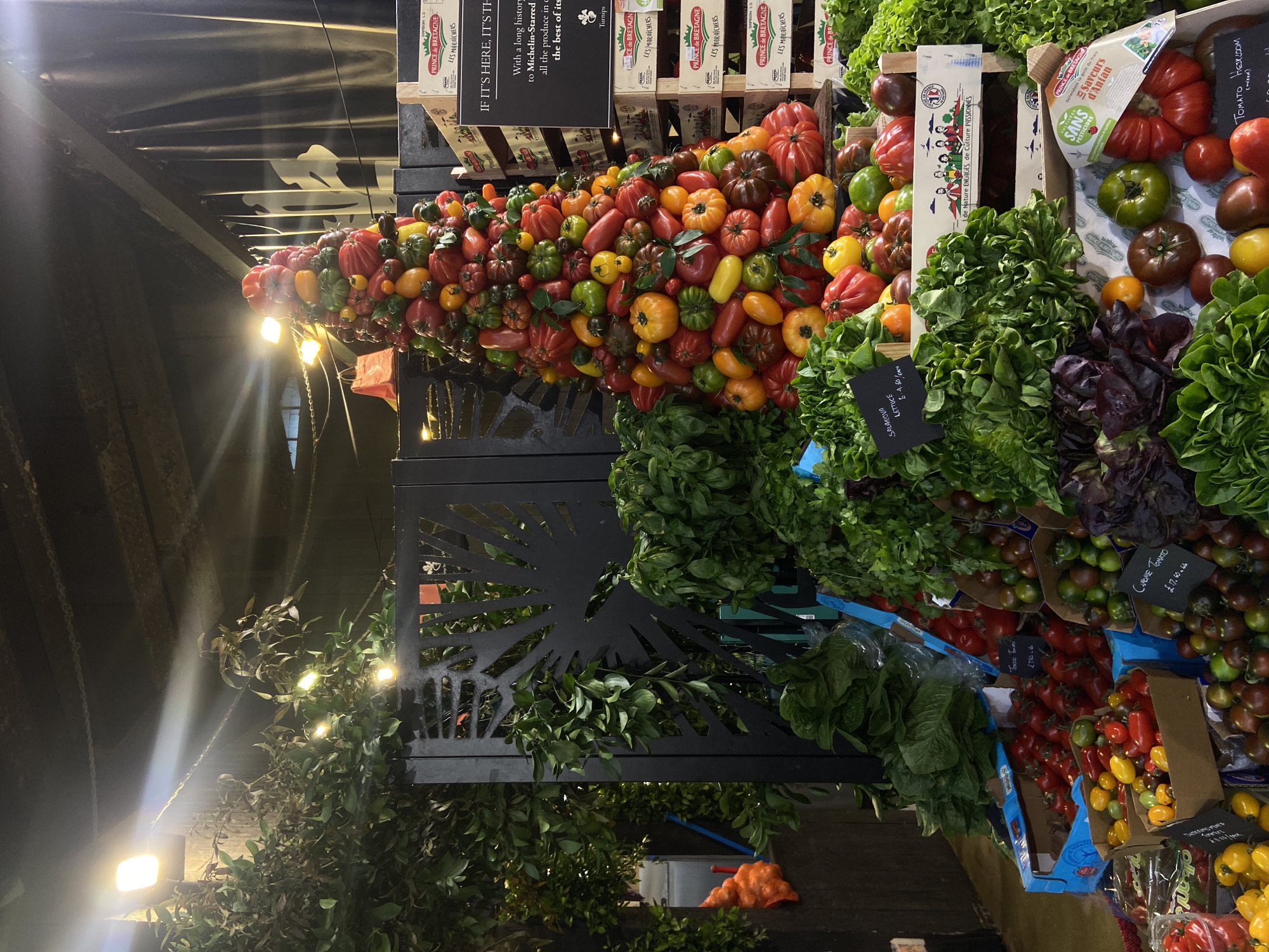 This image captures a vibrant and artfully arranged display of fresh produce, likely within a high-end market stall or grocery store in London, United Kingdom. No people are visible in the frame.
The central subject is a large, conical display of numerous heirloom tomatoes, showcasing a rich variety of colors, shapes, and sizes, including reds, oranges, yellows, greens, and dark purplish-browns. This impressive arrangement is brightly illuminated by artificial lights positioned beneath it.
To the right, various types of leafy greens, predominantly different varieties of lettuce (both green and purple-leafed), are neatly arranged in wooden crates and boxes. Further down, smaller boxes hold cherry tomatoes and other specialty tomato varieties.
Notable visible text includes "PRINCE de BRETAGNE LES MARAICHERS" and "de Nature ENGAGÉS, de Culture PASSIONNÉS" on packaging, indicating the origin and quality commitment of the producers. A label for "Saveurs d'Antan" explicitly states "SANS PESTICIDES" (without pesticides). Specific prices and product names are visible on small chalkboards and labels, such as "TOMATO HEIRLOOM (mixed)," "SALANOVA LETTUCE £6.4.60," "CANOME TOMATO £17.60.6," and "Toma Trio £7.80.4." A larger sign declares, "IF IT'S HERE, IT'S TH With a long histor to Michelin-Starred all the produce in c the best of its," highlighting the premium quality of the produce.
The setting features exposed ceiling beams and dark structural elements in the background, characteristic of an indoor market hall, with additional artificial lights illuminating decorative foliage and metalwork, contributing to a warm, inviting atmosphere.