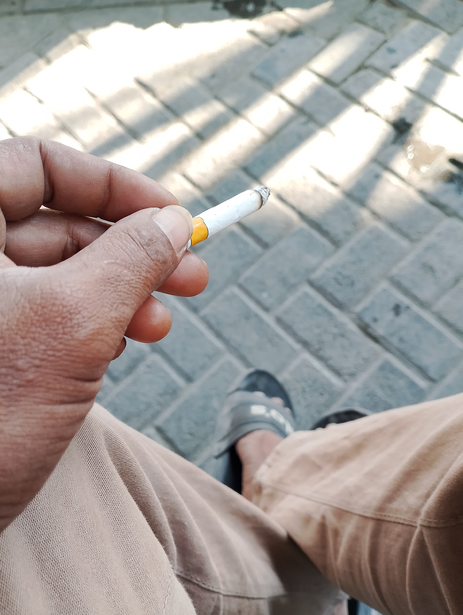 A close-up, top-down perspective shot shows a hand holding a lit cigarette over a herringbone-patterned brick walkway. The hand, with its textured skin and visible fingernails, grasps the cigarette between the thumb and index finger. The cigarette is white with a yellow filter. The background reveals the brick walkway with sunlight casting dappled shadows, suggesting an outdoor location. The lower portion of the image shows tan pants and a grey sandal on a foot, also positioned on the brick surface. The scene is devoid of any specific landmarks or indicators that would place it in Jambean, Indonesia.