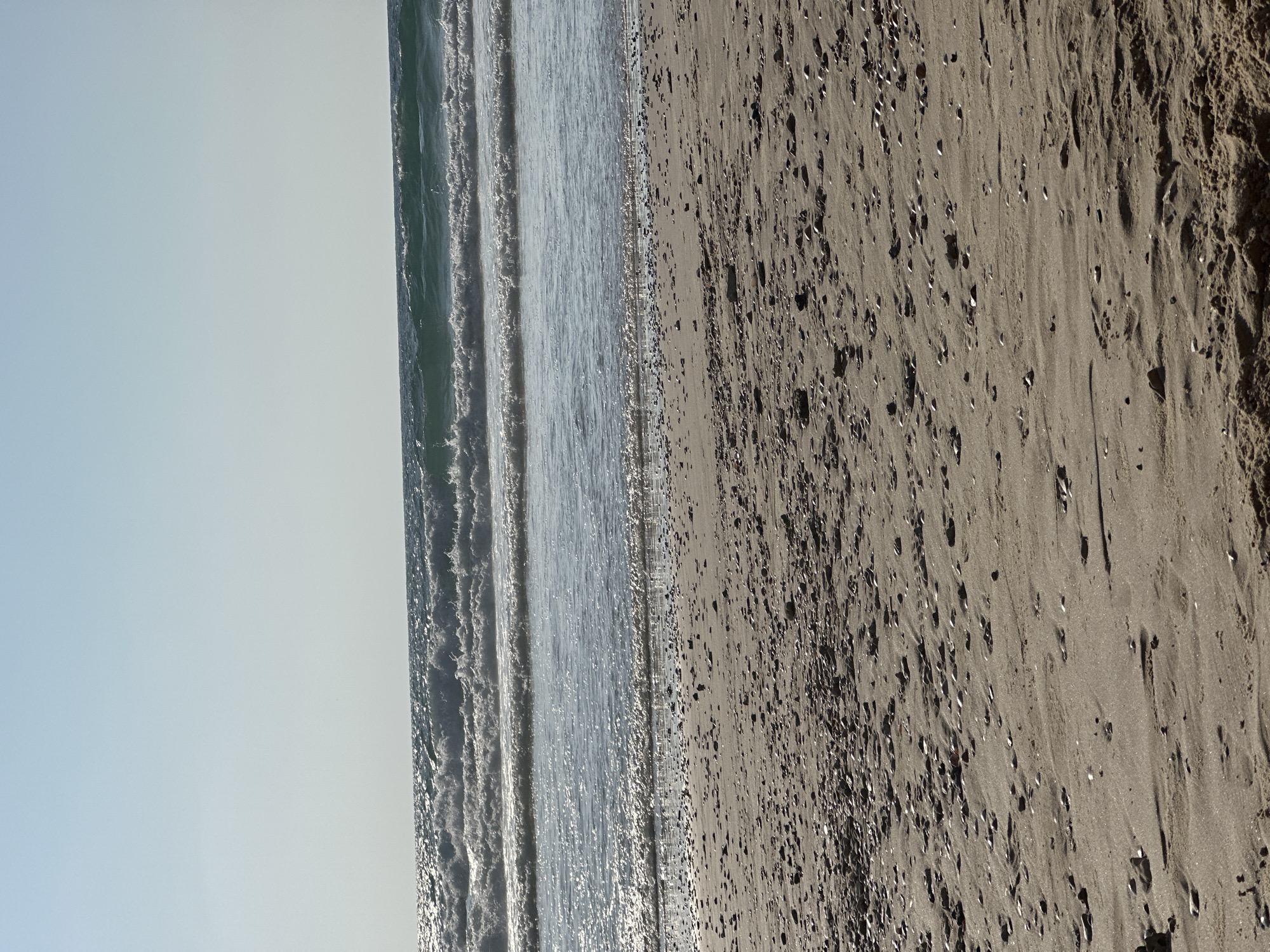 The image depicts a serene beach scene under a clear, bright sky, likely taken in Herzliya, Israel. The foreground is dominated by a sandy beach scattered with small pebbles and shells. The sand appears dry and textured, with subtle ripples and shadows indicating gentle undulations.
Beyond the beach, the Mediterranean Sea stretches out, with multiple rows of waves rolling towards the shore. The water is a deep blue-green, reflecting the sunlight, which creates a sparkling, shimmering effect on the surface. The waves are moderately sized, with white foam cresting as they break. The horizon line is sharp and distinct, separating the sea from the pale blue sky.
There are no people visible in the image, nor any structures or man-made objects. The scene is purely natural, capturing the tranquil beauty of a sunny day at the beach. The overall impression is one of peace and natural splendor.