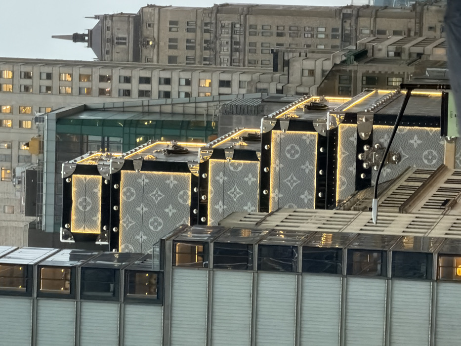 An exterior view in New York City, United States, depicts a building facade transformed into a large-scale architectural installation resembling stacked Louis Vuitton travel trunks. The trunks feature a light grey base adorned with the white Louis Vuitton monogram pattern, accented by black trim, numerous silver-toned metallic rivets, and corner protectors. Warm-toned LED strip lighting outlines each trunk segment and illuminates select interior panels. To the immediate left, a modern building with a facade of light-colored panels and dark windows reflects ambient light. In the background, multiple high-rise buildings are visible, including a prominent light-colored skyscraper with traditional architectural details and a distinct spire-like roof structure. A portion of a black street light pole extends into the lower left foreground. The sky above is overcast. No individuals are visible in the frame.