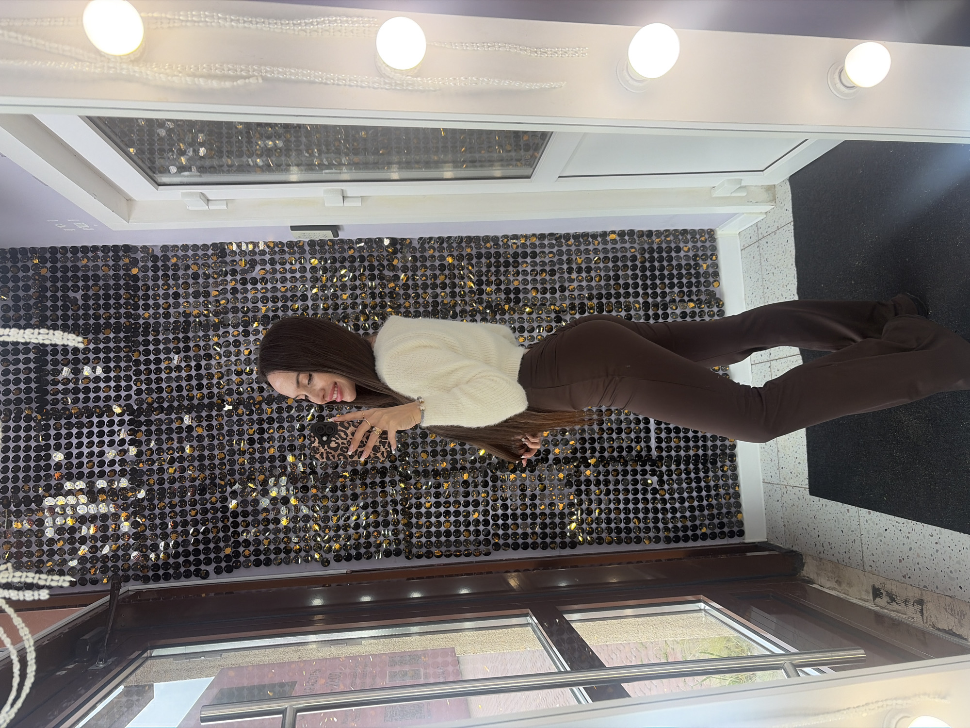 A young woman with long brown hair is standing indoors, posing for a selfie. She is wearing a fluffy white sweater and brown bell-bottom pants. The background features a wall covered in black and gold sequins, and to her right is a white door with a window. To the left of the woman is a large window with a dark brown frame, and beyond the window, a glimpse of a street with buildings can be seen. The floor is tiled with a speckled pattern. In the foreground, a black mat is visible. The lighting appears to be artificial, possibly from nearby lights or a ring light. The scene is likely in a dressing room or a public space with a decorative wall. There is no information in the image to indicate a specific location in Saint Petersburg, Russia.