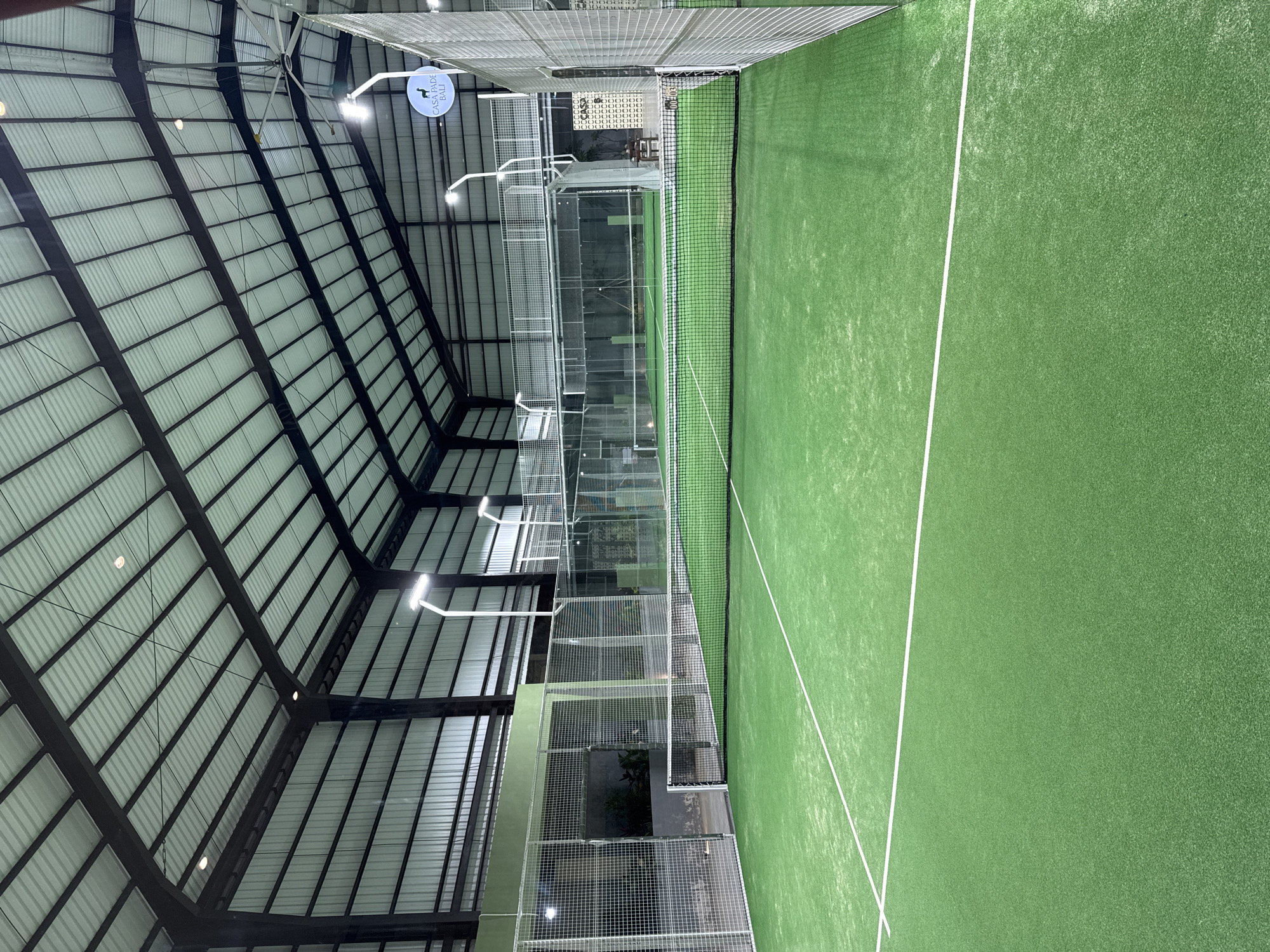 The image depicts an indoor padel court at Casa Padel Bali in Kuta Selatan, Indonesia. The court has a green surface and white lines. A net spans the center. Wire mesh surrounds the court. The roof structure is metal with support beams. Artificial lighting is visible.
