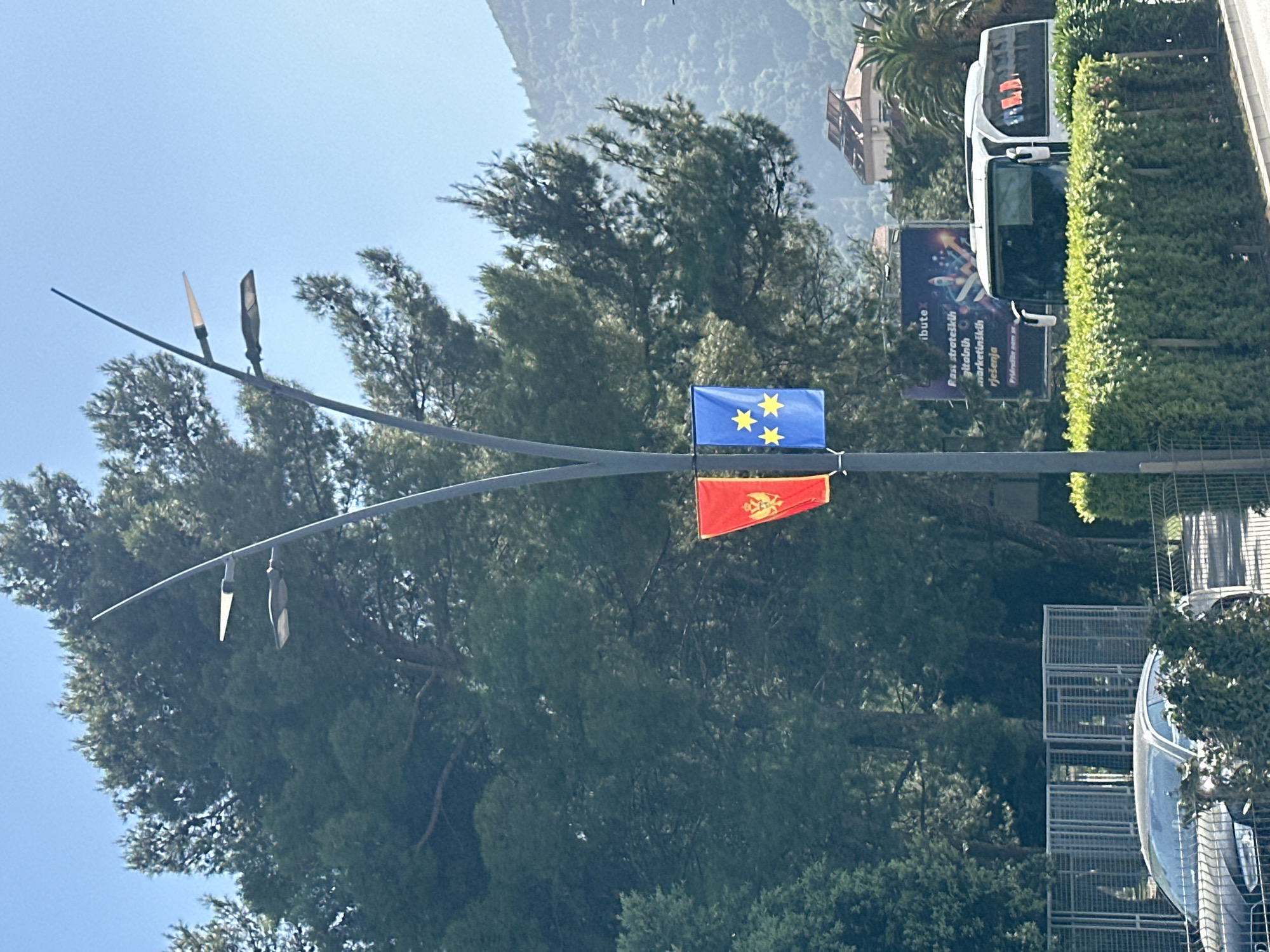 The image captures a street scene in Budva, Montenegro. A metal street lamp is in the foreground, with the European Union flag and the Montenegrin flag attached to it. The EU flag is blue with three yellow stars, while the Montenegrin flag is red with a gold coat of arms. Behind the lamp post, dense green trees fill much of the frame. Further back, parts of buildings and a mountain are visible. A large white bus is parked on the right side of the image, partially obscured by a green hedge. A billboard with the word "ibute" and some graphics is also visible near the bus. Below, to the right, a silver car and metal fencing are partially visible. The sky is light blue, suggesting a clear day.