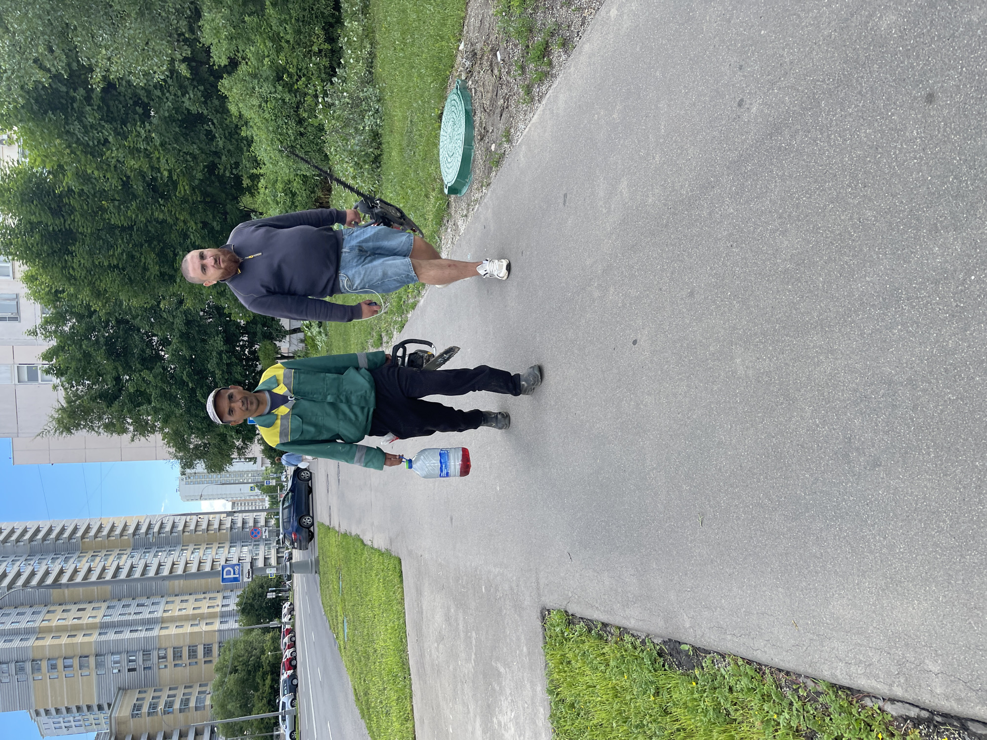 Two men are walking on a paved sidewalk in Moscow, Russia. The man on the left, wearing a green and yellow work jacket and black pants, is carrying a large plastic bottle filled with a red liquid in his right hand. He is also wearing a white baseball cap. The man on the right, wearing a dark blue sweatshirt and denim shorts, is carrying a chainsaw horizontally in his right hand. He has a beard and is wearing white sneakers. They are walking away from the camera and towards a grassy area with bushes and trees. In the background, there are tall apartment buildings and a street with cars. A green manhole cover is visible on the grass to the right of the path. The sky is blue with some clouds.