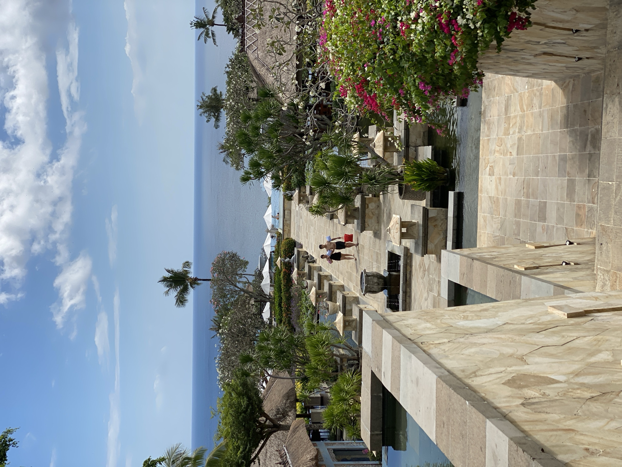 The image depicts a resort in Kuta Selatan, Indonesia, overlooking the ocean. A paved pathway, lined with decorative urns and planters, leads down towards the water. Two individuals, a man and a woman, are walking along the pathway, carrying towels. Lush green vegetation, including palm trees and flowering bushes, surrounds the area. White umbrellas are visible near a pool in the background, and traditional thatched-roof buildings are scattered throughout the resort. The sky is clear with scattered white clouds.