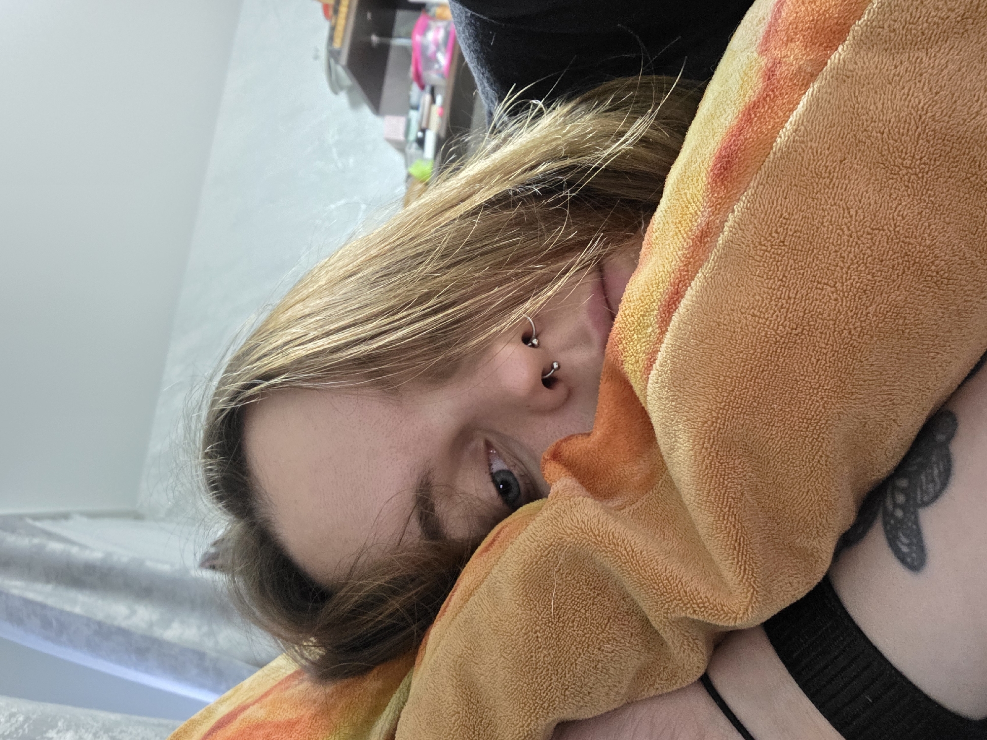 A close-up, tilted portrait shows a person partially obscured by an orange and yellow towel. The person has fair skin, light brown hair, and blue eyes. Visible facial features include an eye, part of the nose with a piercing, and a glimpse of the mouth. A black article of clothing is visible above the head and also near the shoulder. A tattoo, likely of a butterfly or similar design, is visible on the person's shoulder. The background includes a white wall and glimpses of furniture. The location is specified as Unknown, Unknown.