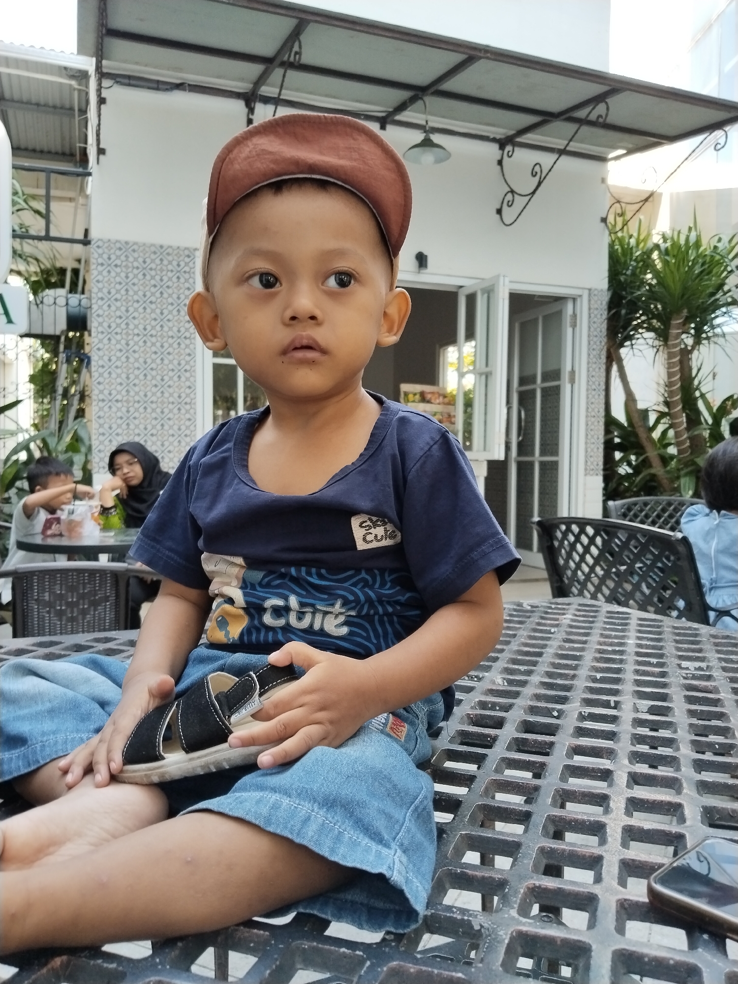 A young boy sits at a table, his gaze directed off-camera to the right. He wears a navy blue t-shirt with a blue and white patterned design, denim jeans, and a brown baseball cap worn backwards. In his hands, he holds a black sandal with white trim. The boy's expression appears neutral, with his mouth slightly open.
The setting is outdoors, likely a cafe or restaurant, given the tables and chairs visible. The tables have a black, grid-like metal surface. In the background, a building with white walls and open doors is visible, along with plants and trees. Other people are seated at tables in the background, including a woman wearing a hijab and a child. The lighting suggests it is daytime. The overall atmosphere seems relaxed and casual.
A cell phone is visible on the table in the lower right corner of the frame. The text "Cute" is visible on the boy's t-shirt.