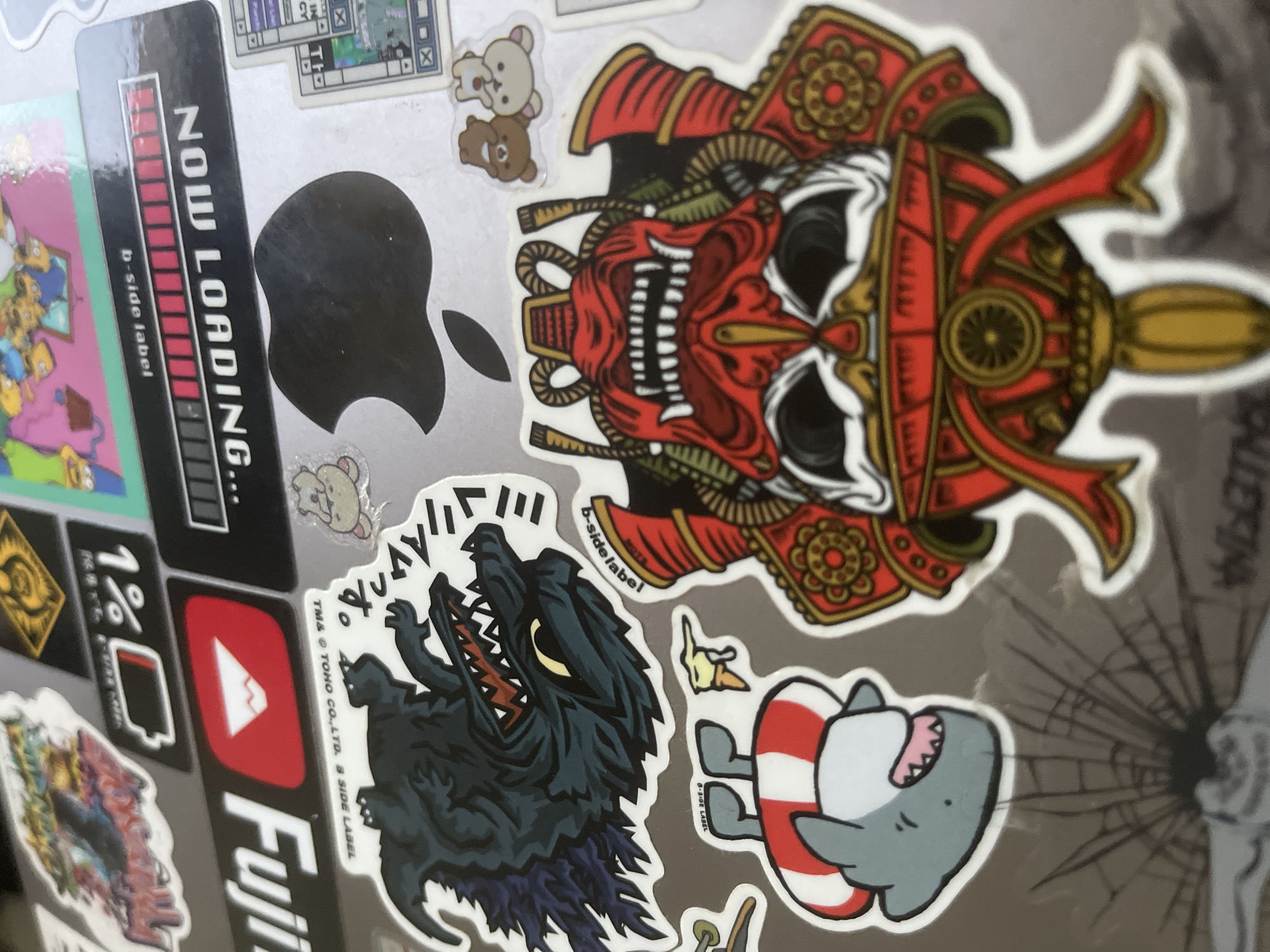 This is a close-up, high-angle shot of a laptop surface covered in various stickers. The stickers feature a range of designs, including a stylized wolf or monster, a loading bar graphic with the text "NOW LOADING.", a YouTube logo, two small cartoon bears, a cartoon shark wearing a life preserver, and a Japanese-inspired samurai mask. The stickers are colorful and overlap in some areas. The laptop surface itself appears to be metallic, possibly silver or gray. The lighting suggests an indoor setting. There are no people or any elements that suggest a location in Paris, France.