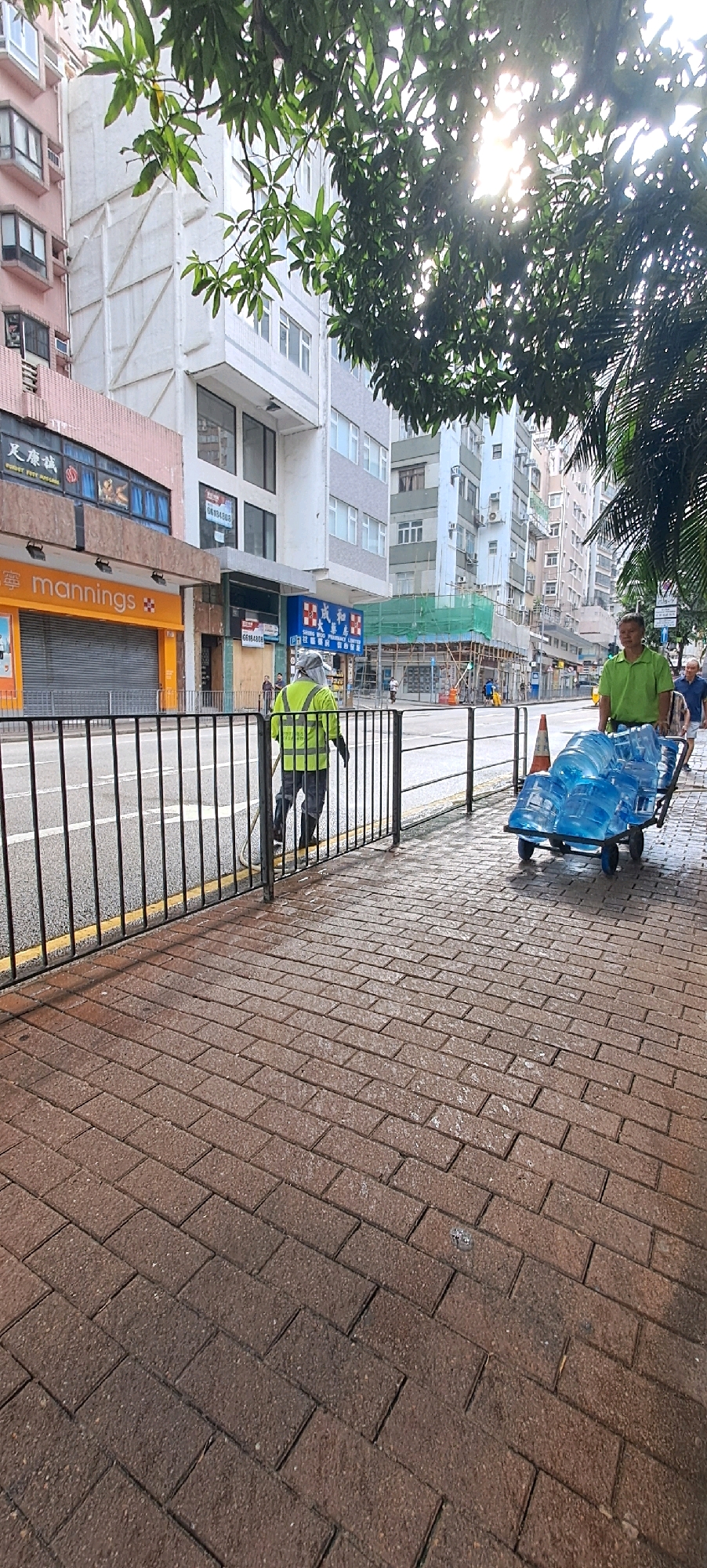 The image captures a street scene in Hong Kong, China. In the foreground, a person wearing a high-visibility yellow vest and a face covering is sweeping the brick-paved sidewalk with a broom. They are positioned behind a black metal fence that separates the sidewalk from the street. To the right of the sweeper, a man in a green polo shirt is pushing a cart laden with large, blue water cooler bottles. The cart has two wheels and a handle. An orange traffic cone is visible near the cart.
The background shows a typical urban environment with multi-story buildings lining the street. The buildings have various architectural styles and colors, including pink, white, and grey. Signs are visible on some of the buildings, including one that reads "mannings" in orange lettering. Construction scaffolding covered with green netting is present on one of the buildings. Sunlight filters through the leaves of a tree in the upper part of the image, casting dappled light on the scene. The street itself appears wet, suggesting recent rain or street cleaning. The overall impression is one of an active street with people engaged in their daily tasks.