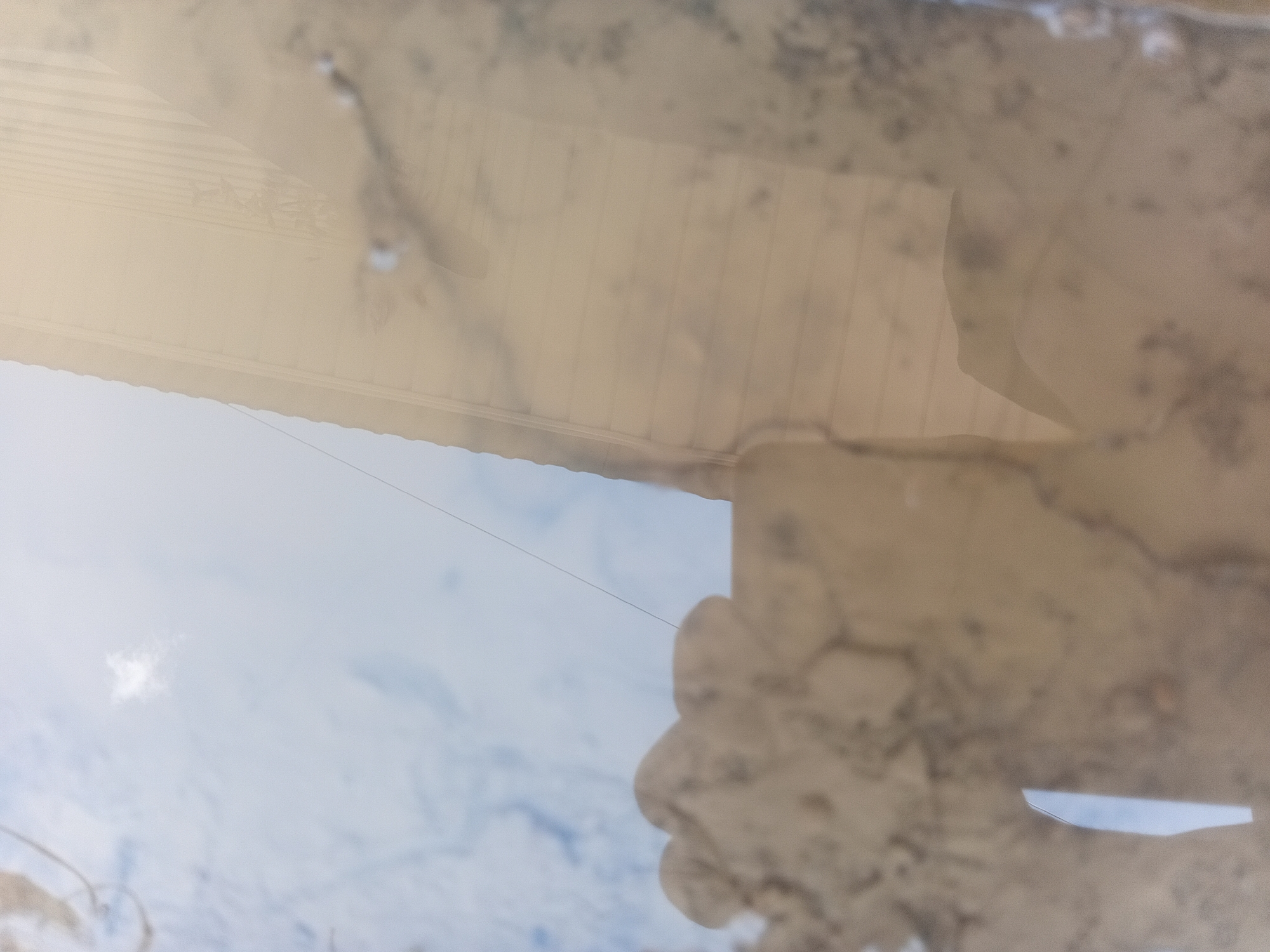 A vertical, close-up, abstract shot of a reflective surface, possibly water, shows a distorted reflection of the sky and a building's exterior. The sky appears as a pale blue with scattered clouds. The building's reflection is a light tan with faint, horizontal lines, suggesting siding or paneling. The texture of the reflective surface is visible, with some mottled patterns and a bright, vertical streak that could be a reflection of a light source or an object. The scene appears to be outdoors, with the sky being a dominant element in the reflection. The location within Verkhniachka, Ukraine is not discernible from the image itself.