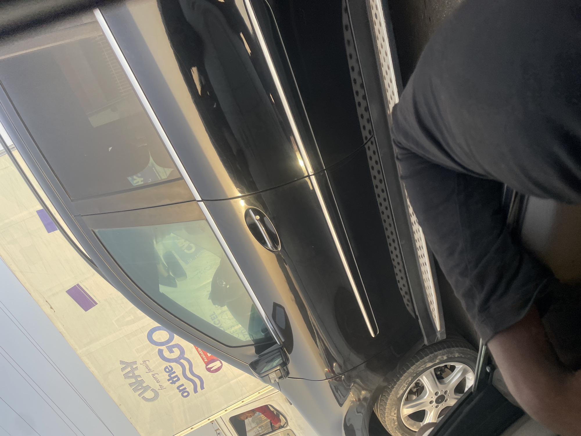 The image displays a tilted, close-up view of a dark-colored SUV, likely black, under bright daylight conditions in Abuja, Nigeria. The vehicle's side is visible, featuring a prominent chrome door handle, chrome trim around the windows, and a silver, textured side running board. Part of a multi-spoke silver wheel rim and tire is also seen in the lower right.
A person, wearing a dark short-sleeved top, is partially visible in the bottom right, appearing to be engaged with the vehicle, possibly entering or leaning into it. Through the SUV's tinted windows, hints of the interior are discernible, along with reflections.
The glossy surface of the SUV strongly reflects its surroundings. A large white vehicle, likely a truck or van, is prominently reflected on its side. This reflected vehicle carries advertising with clear text: "CWAY" and "on the GO," with the tagline "for every family" underneath "CWAY." Other fainter text like "Available" and possibly "EXCEL" is also visible in the reflections. The bright reflections and highlights on the SUV indicate strong sunlight, suggesting a clear, sunny day. Faint power lines are visible against the bright sky in the upper left background. The overall scene captures a moment of everyday activity involving vehicles.