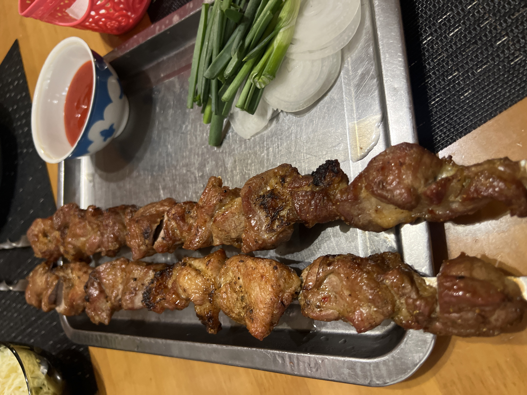 A high-angle medium shot depicts a metal serving tray containing two skewers of grilled meat, green onions, sliced onions, and a small bowl of red sauce. The tray sits on a wooden surface. A red plastic basket is partially visible in the upper-left corner, and a container of a yellow substance is partly visible in the lower-left corner.