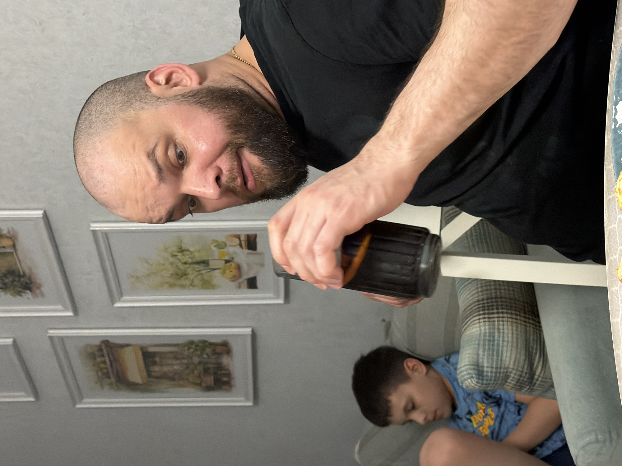 An indoor scene captures a man and a young boy, with the man in the foreground and the boy resting in the background. The man, with a shaved head and a beard, is wearing a black t-shirt and a thin gold chain. He is looking slightly downwards and to his right, with a somewhat serious or contemplative expression. His left hand holds a dark-colored bottle, possibly of a beverage like beer or soda, which he appears to be either holding up or pouring from, though the action isn't fully clear.
In the background, to the right and slightly below the man, a young boy with short, dark hair is seen sleeping or resting. He is wearing a blue t-shirt with a yellow graphic design and is lying on a light-colored sofa or cushioned surface, partially obscured by a striped pillow and a white furniture piece (possibly a table leg or chair arm).
The background wall is light gray, adorned with several framed pictures, possibly depicting landscapes or still life art, though the details within the frames are blurry. The overall setting suggests a casual home environment. No specific time of day is indicated, given the indoor lighting. There is no visible text in the image.