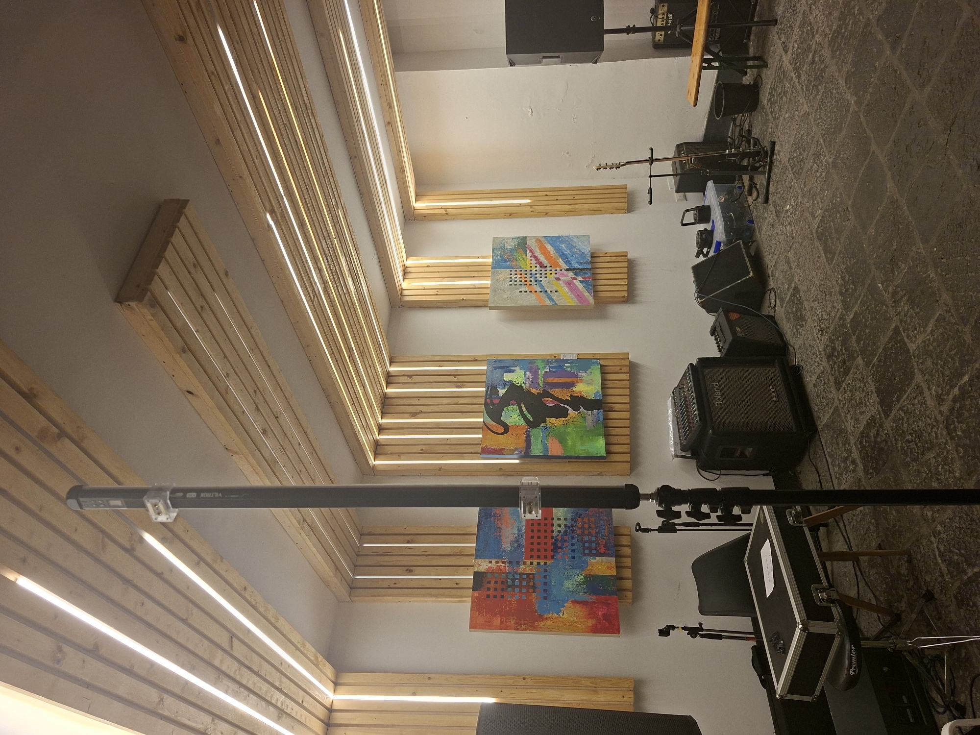 The image depicts an interior space, likely a studio or rehearsal room, in Messina, Italy. The walls are white with horizontal wooden slats, and linear lights are positioned between the slats, creating a modern aesthetic. Three framed abstract paintings adorn the wall. Musical equipment, including a guitar, amplifier, and a keyboard setup with a chair, are scattered around the room. The floor is paved with dark stones. A black boom pole with a microphone attached is extended across the room. The scene appears to be set up for music recording or practice.