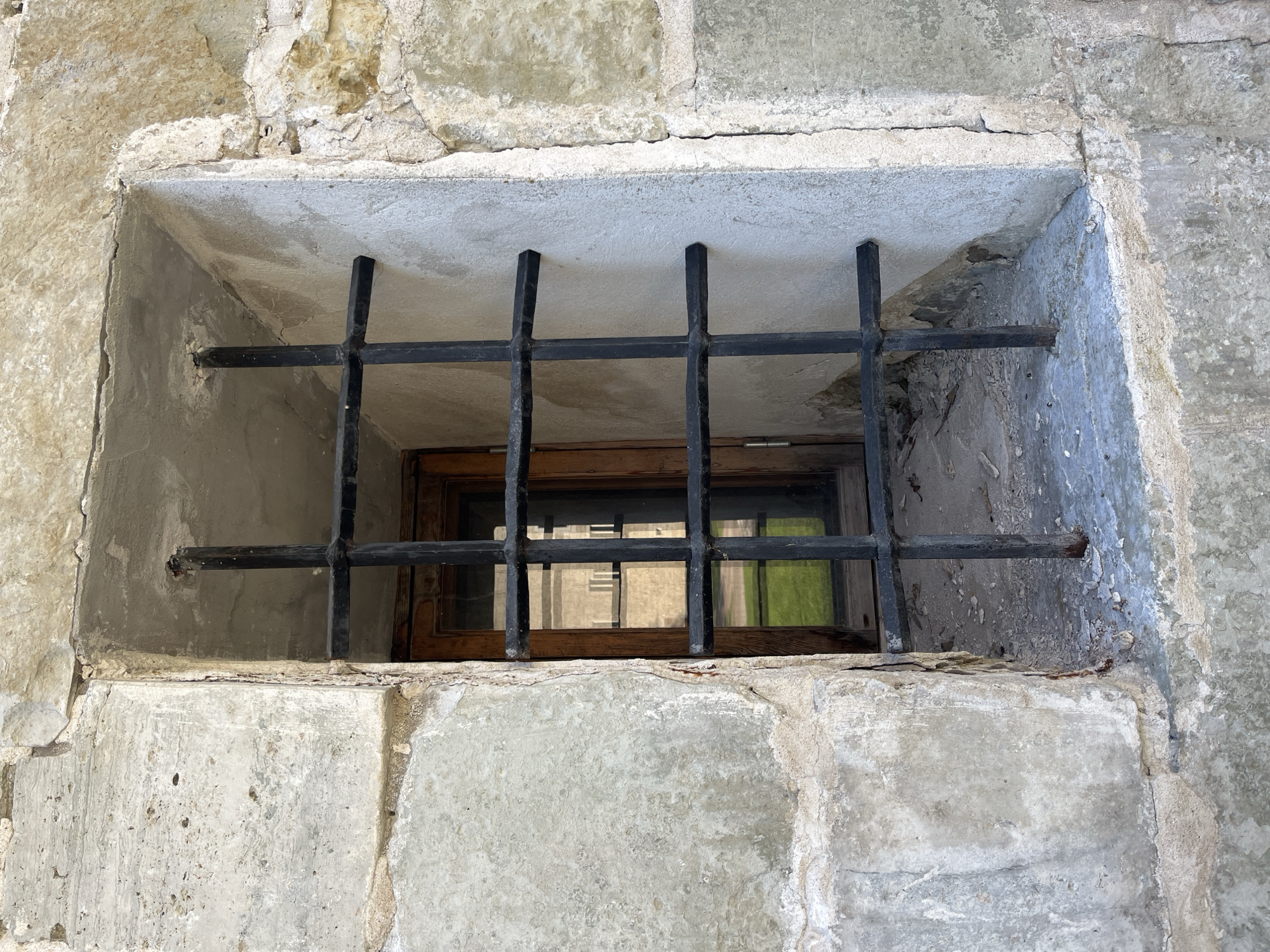 This is a close-up, eye-level, medium shot of a barred window in an old stone building. The window is recessed into the wall, which is constructed of large, rough-hewn stone blocks. The bars are made of thick, black metal and are arranged in a grid pattern. The window frame appears to be made of wood, and through the glass, a reflection of another building and a patch of green grass is visible. The sill of the window is covered in dust and debris. The overall impression is one of age and perhaps neglect. The building is likely a historical structure, possibly a prison or a castle.
Based on the architectural style and the presence of the specific window design, this image is likely from Kuressaare Castle, also known as Kuressaare Episcopal Castle, located in Kuressaare, Estonia. The castle is a well-preserved medieval fortress with a history dating back to the 14th century.