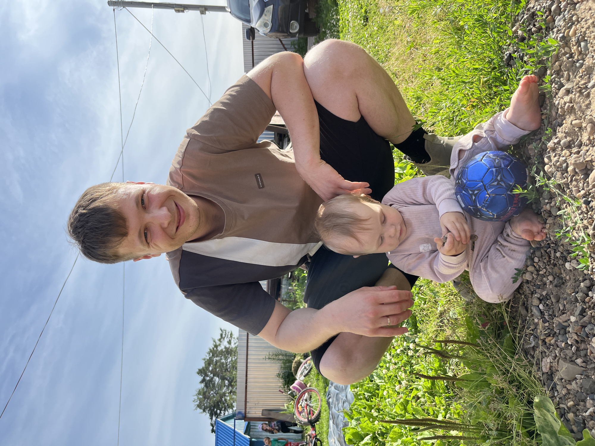 A man and a baby are outdoors in what appears to be a rural setting, likely in Chaikovsky, Russia, given the context of the request. The man is squatting and looking at the camera with a smile, wearing a two-toned t-shirt and black shorts. In front of him, the baby is sitting on the ground playing with small stones, dressed in a light pink onesie. A blue soccer ball rests beside the baby. The background shows a fenced yard with a corrugated metal fence, a blue building, trees, a bicycle, and a car. The ground is covered with grass and gravel. The sky is blue with scattered clouds, suggesting daytime.