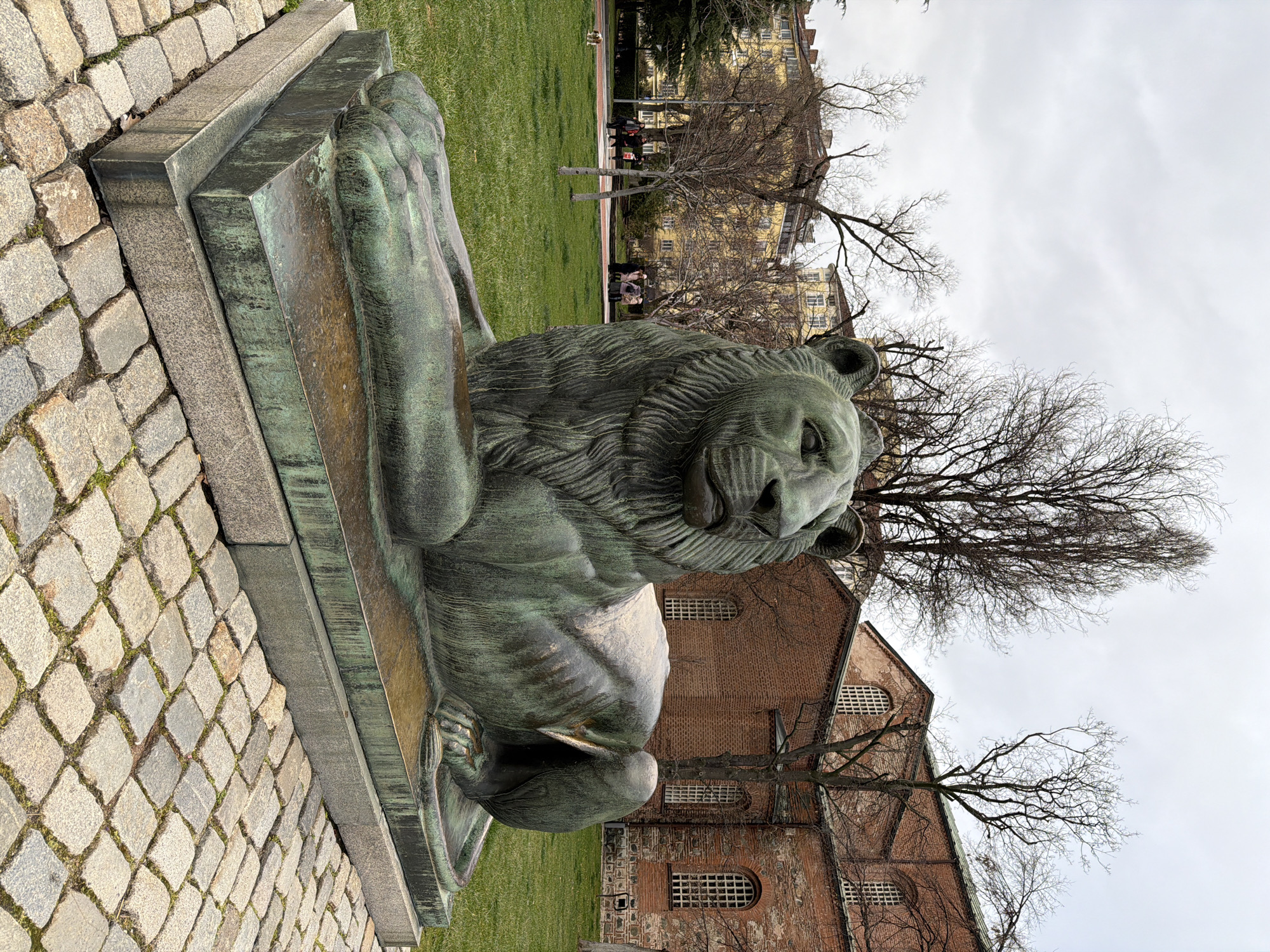 The image captures a statue of a recumbent lion, crafted from a green-tinged metal, situated on a stone base, with a cobblestone ground in the foreground. The background reveals a grassy park area with trees, some bare and others starting to show signs of foliage. Buildings with brick facades and windowed details are partially visible behind the trees, suggesting an urban setting. The scene is under an overcast sky, casting a muted light on the various elements. The lion statue is the prominent subject. There are no distinct people visible in the foreground, but the background shows potential figures walking or sitting in the park. The interaction primarily involves the statue being positioned in a public park, set against the architectural backdrop. The location is the city of Unknown, Unknown.