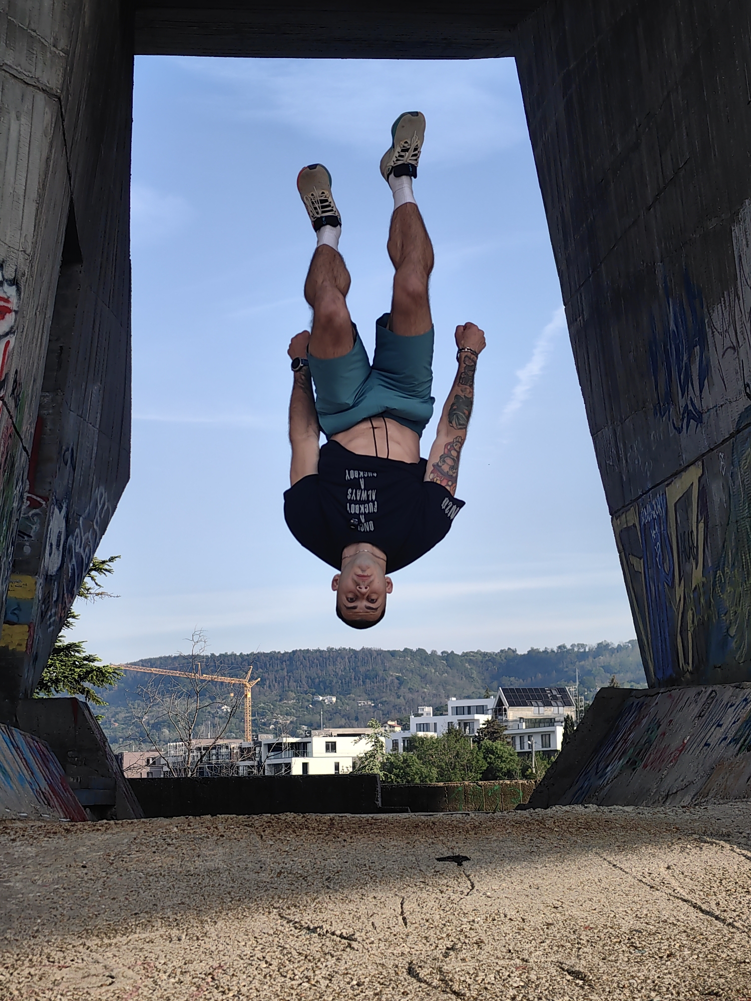 A man is performing a handstand in an urban environment. He is upside down, facing the camera, with his legs extended upward and his hands clenched into fists at his sides. He is wearing a black t-shirt, green shorts, white socks, and tan-colored sneakers. His skin is fair, and he has dark hair. Tattoos are visible on his forearms. The background is an open structure of concrete walls and roof, framing a view of a city with white buildings, green trees, and a distant hill with a construction crane. The sky is blue with wispy clouds. The ground beneath the man appears to be textured concrete. The image is taken in Unknown, Unknown.