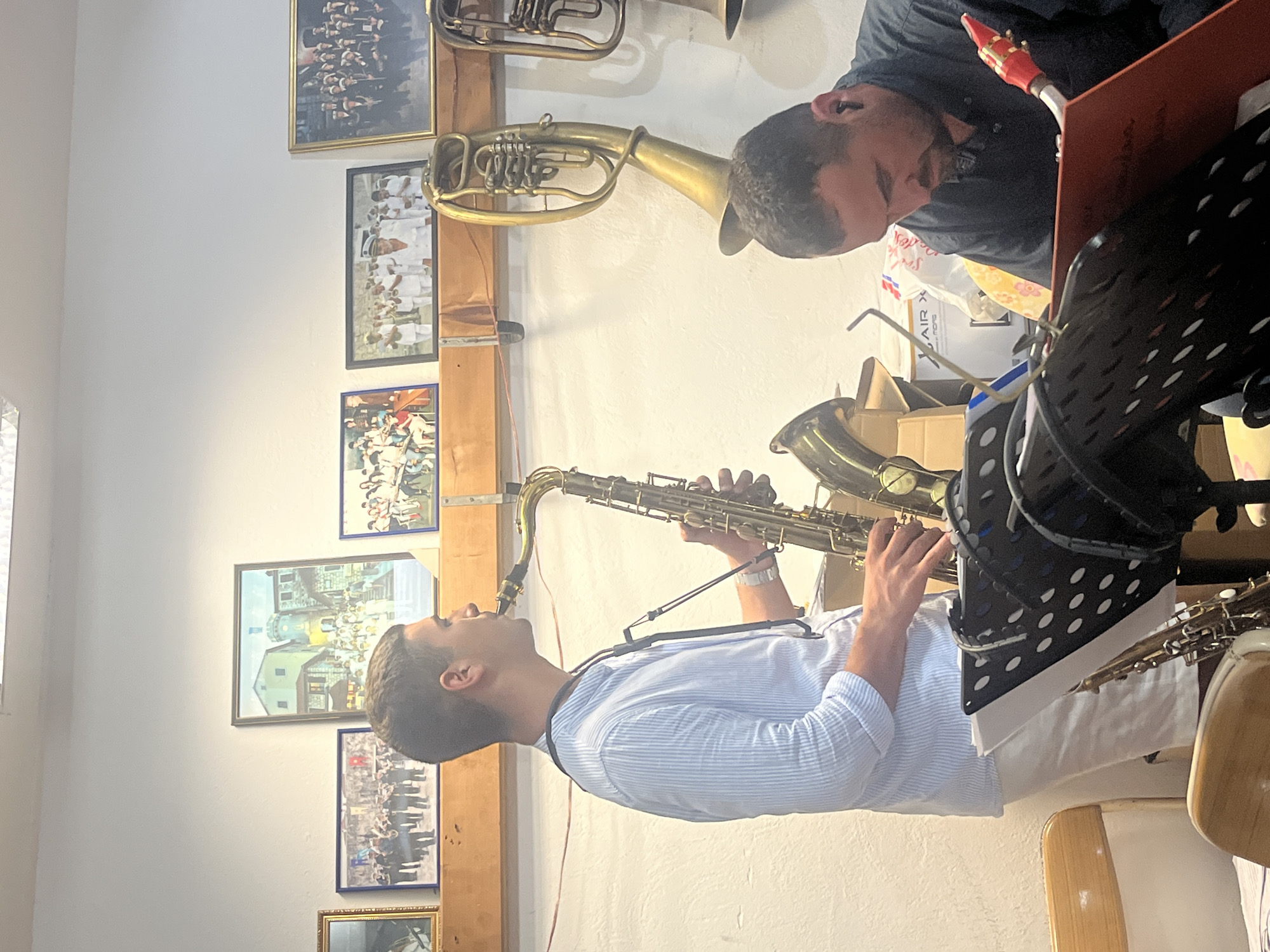 In an interior space with white walls and a wooden shelf, two men are present. One man, on the left, is playing a tenor saxophone. He is wearing a light blue striped, collared shirt and white shorts. He has a watch on his left wrist and a black strap for the saxophone around his neck. The other man, on the right, is looking down at a red music book and appears to be wearing in-ear monitors. He has dark hair and a beard and is wearing a dark colored shirt. Various framed photographs of groups of people and buildings hang on the wall behind the men. Additionally, several brass instruments, including a euphonium, are visible, some hanging from the shelf. In the foreground, two music stands are present, one with sheet music. The scene appears to be in Budva, Montenegro, possibly in a music studio or practice space.