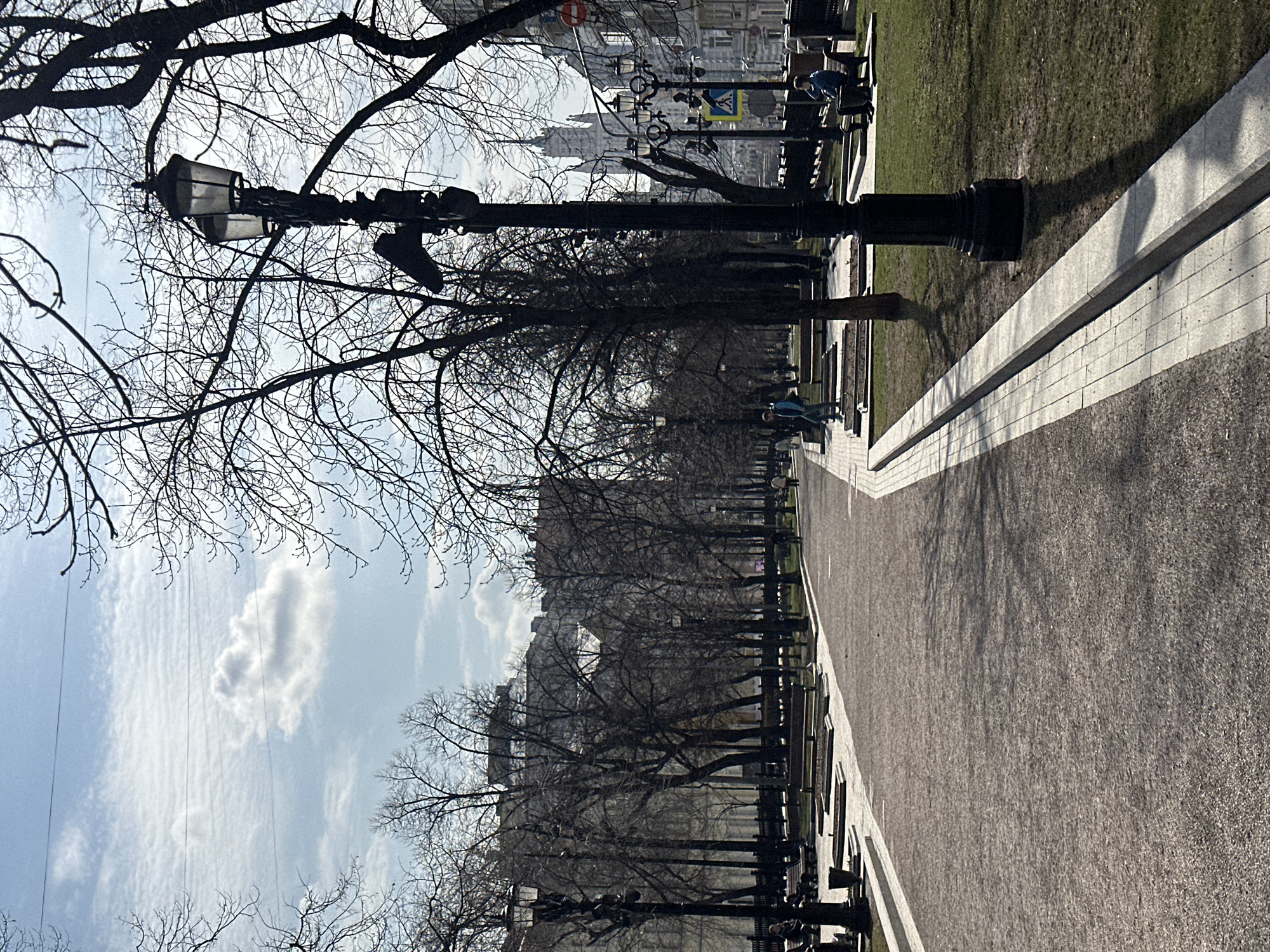 The image depicts a walkway in a park setting in Moscow, Russia, flanked by rows of bare trees and antique lampposts. The walkway is paved with a gravel-like material and bordered by a white stone edge. Benches are positioned alongside the walkway, some occupied by individuals. Buildings are visible in the background through the trees. The sky is partially cloudy. The lampposts are black with ornate detailing.