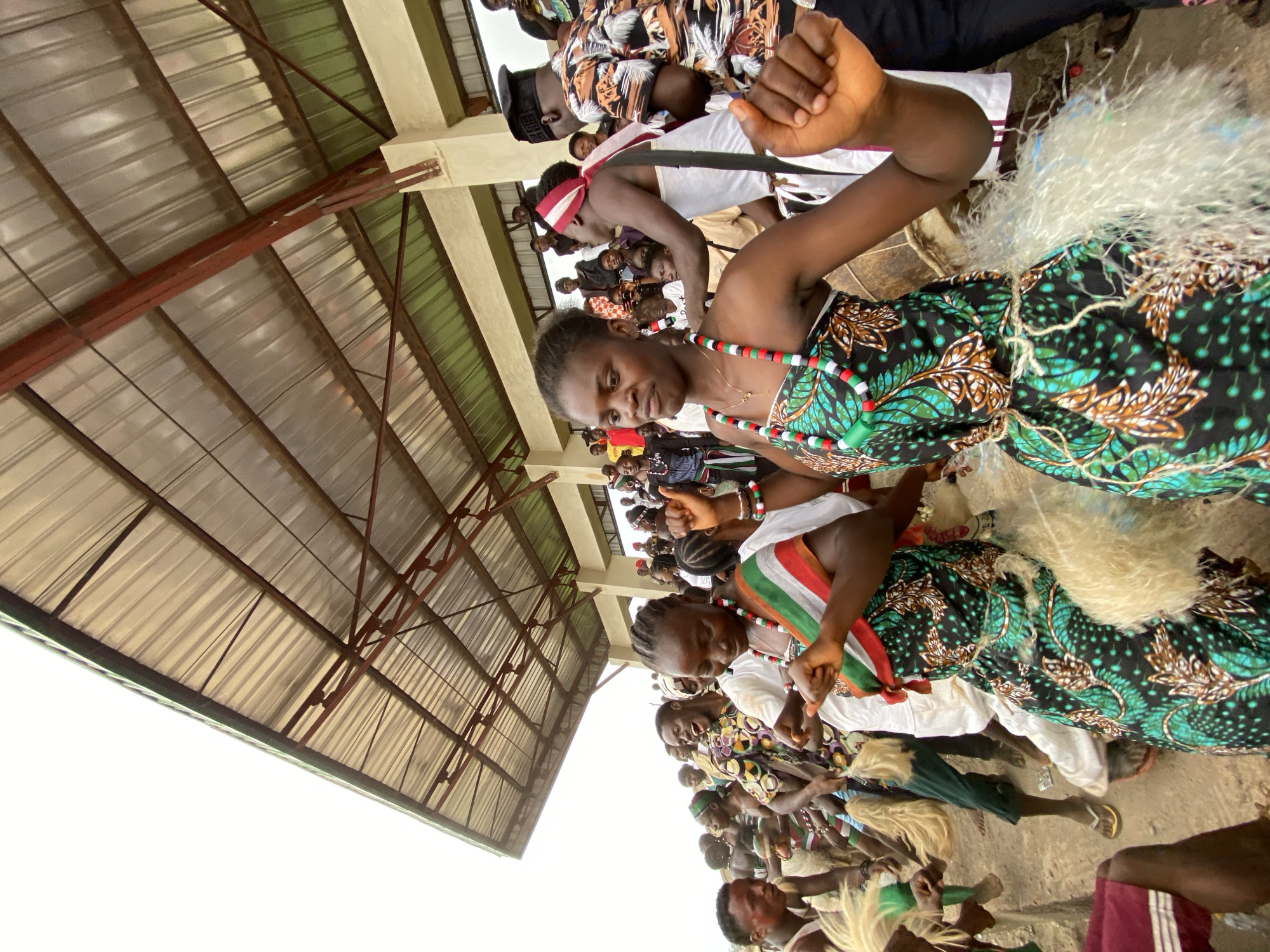 A group of people is gathered under a corrugated metal roof structure supported by steel beams and concrete pillars. The individuals are dressed in colorful, patterned clothing and bead necklaces, suggesting a cultural event. Some people are wearing white garments, while others have white furry attachments around their waists. Some people are holding green and white fabric strips, and some are looking at the camera. The location is Durimi, Nigeria.