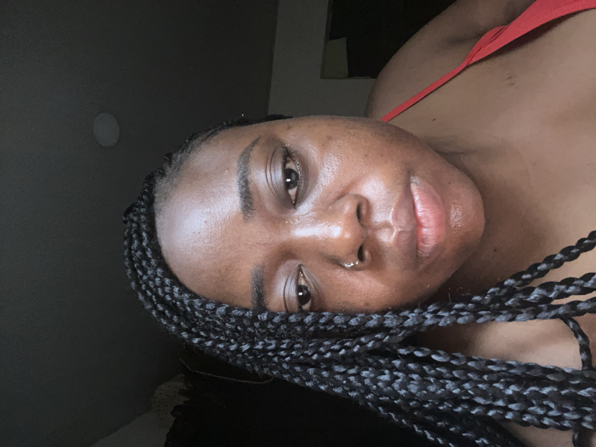 A close-up, tilted portrait of a Black woman with dark braids, potentially indoors with dim lighting. She has a nose piercing. Part of a red strap or garment is visible on her shoulder. The location in Abuja, Nigeria, is not discernible from the image.
