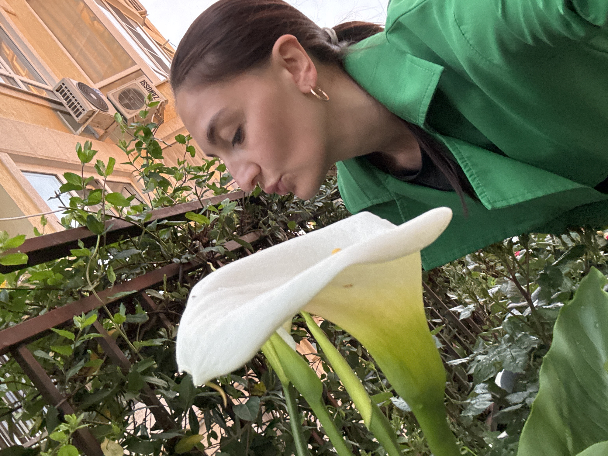 A low-angle shot captures a fair-skinned woman in a green jacket, leaning down to seemingly smell a large, white calla lily. Her hair is pulled back, revealing a gold hoop earring. The background features a building with air conditioning units mounted on its exterior, and a rusty metal fence partially obscured by dense green foliage. The sky is overcast, and based on the architectural style and plant life, the location is likely in Sochi, Russia.
