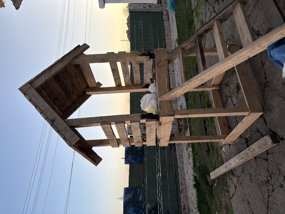 The scene depicts an outdoor residential area in Tarnava, Bulgaria, during late afternoon or early evening. A partially constructed elevated wooden structure stands centrally. The structure is built from rough-hewn lumber, featuring an open base frame, an elevated platform with partial slatted walls, and a gabled roof. A wooden staircase is affixed, providing access to the platform. On the platform surface, a white plastic bag containing unknown contents and several black and orange clamps are visible. The immediate ground consists of cracked concrete pavement mixed with patches of grass. A loose wooden plank lies horizontally on the concrete to the left of the structure. In the midground, a green corrugated metal fence with a stone base extends horizontally. Beyond the fence, a multi-tiered green structure topped with panels featuring blue abstract patterns is present. Utility lines traverse the upper left sky, which transitions from clear blue to an orange-yellow hue towards the western horizon. No individuals are present in the image.