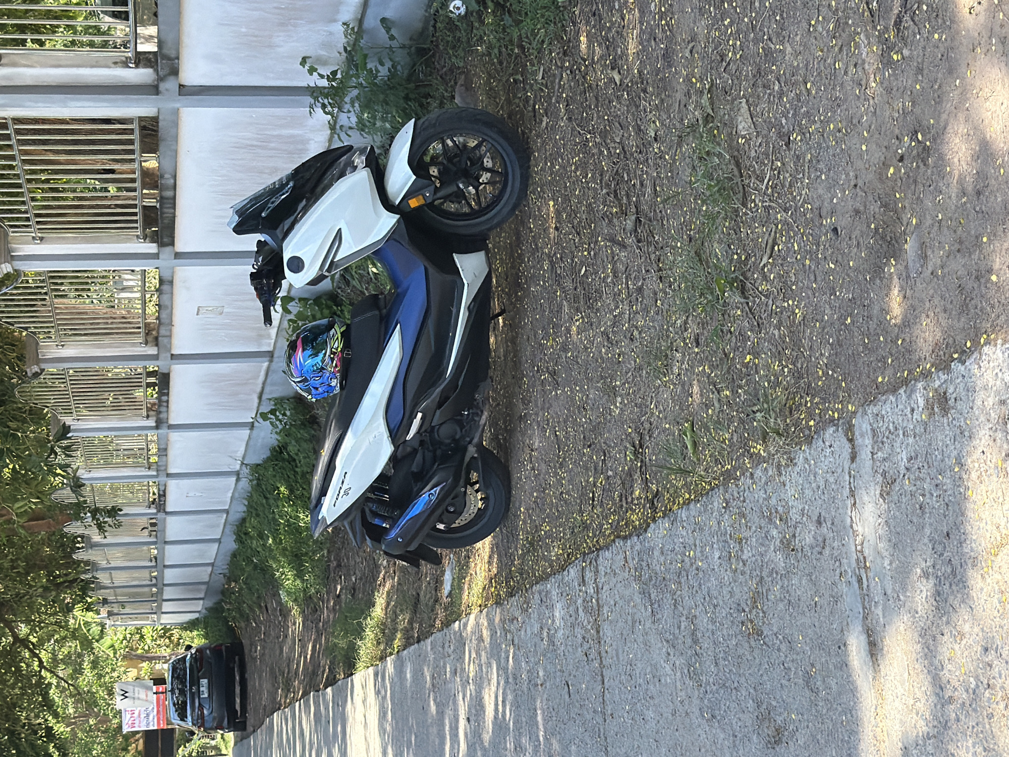 A white and blue scooter is parked on a dirt path next to a concrete walkway. A black helmet with a blue and white design is placed on the scooter's seat. In the background, a dark-colored sedan is parked on the side of the road. A white concrete wall with a metal fence runs along the edge of the property, bordered by trees and vegetation. The scene appears to be in a semi-urban area in Baan Bo Phut, Thailand, given the presence of the car, scooter, and residential wall. The ground is covered with dirt, fallen yellow petals, and some dry leaves and twigs. Sunlight casts shadows from the trees and the walkway onto the ground.